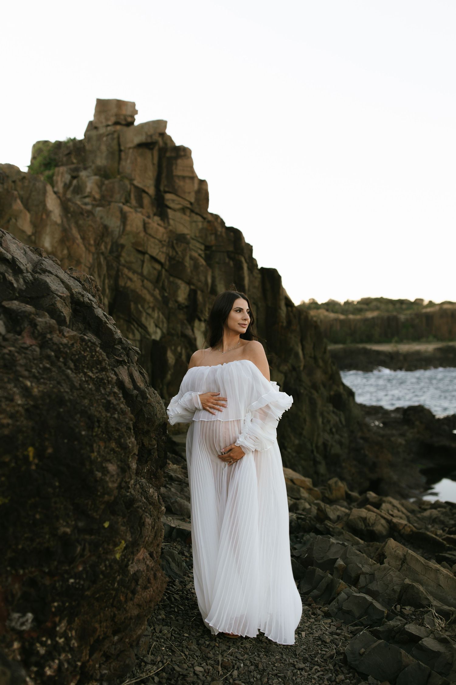 Figure in a flowing white dress stands on rocky coastal terrain against dramatic ocean cliffs at sunset.
