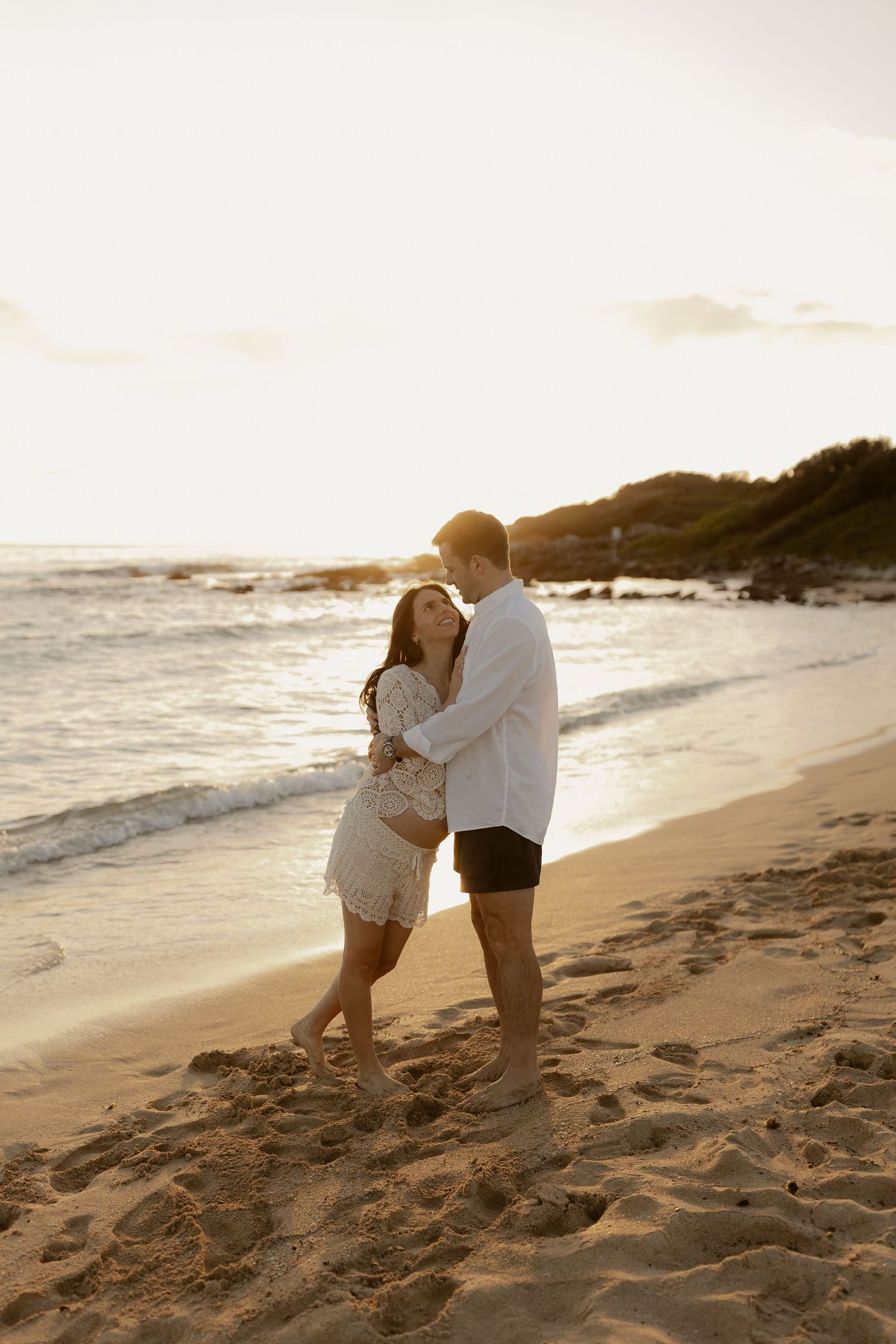 A romantic beach photoshoot at sunset with a couple in white clothing embracing on the sandy shore.