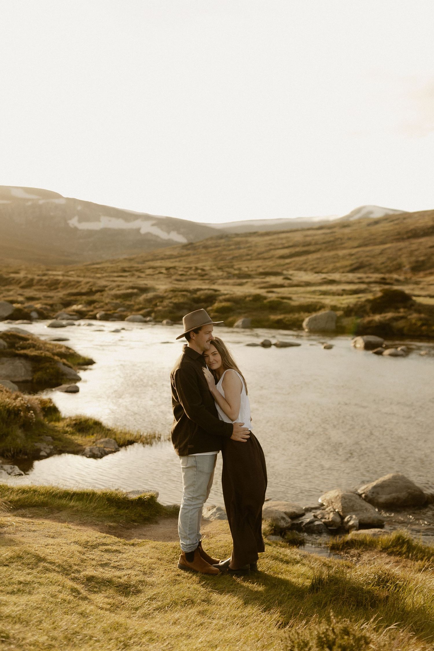 Two people stand together overlooking a serene lake in the Scottish highlands during golden hour.