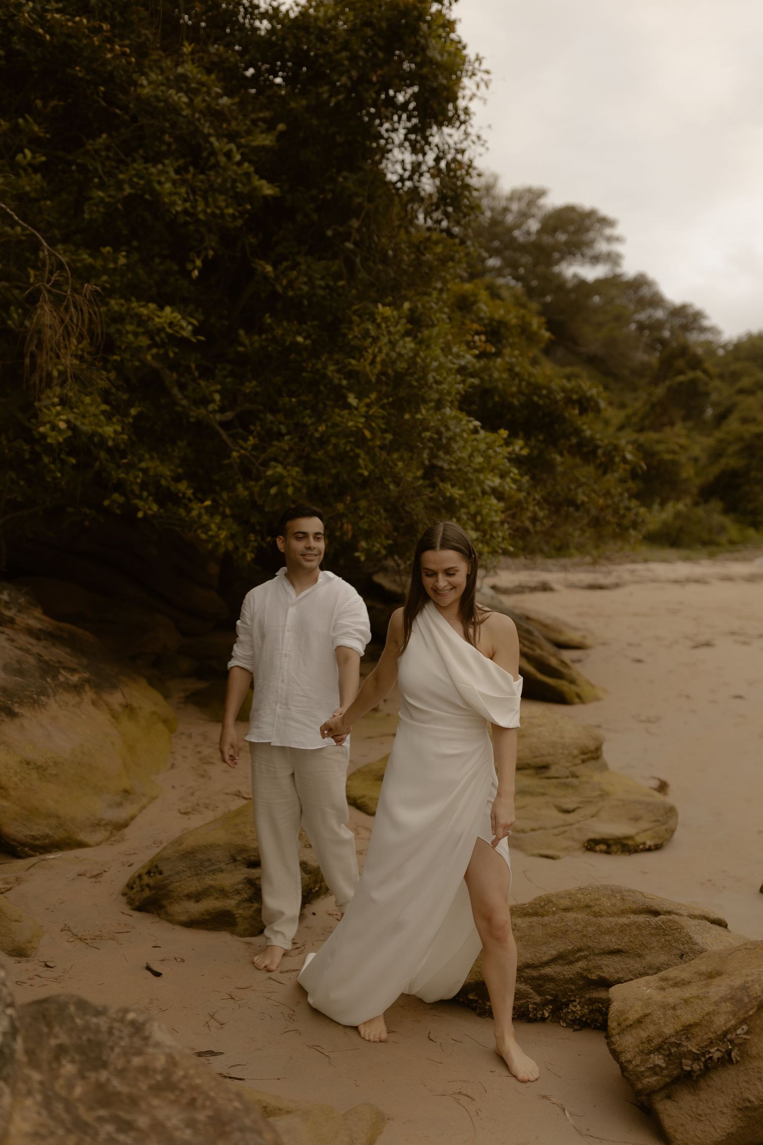 A couple in white outfits sits together on rocky coastal terrain during a romantic photoshoot at sunset.