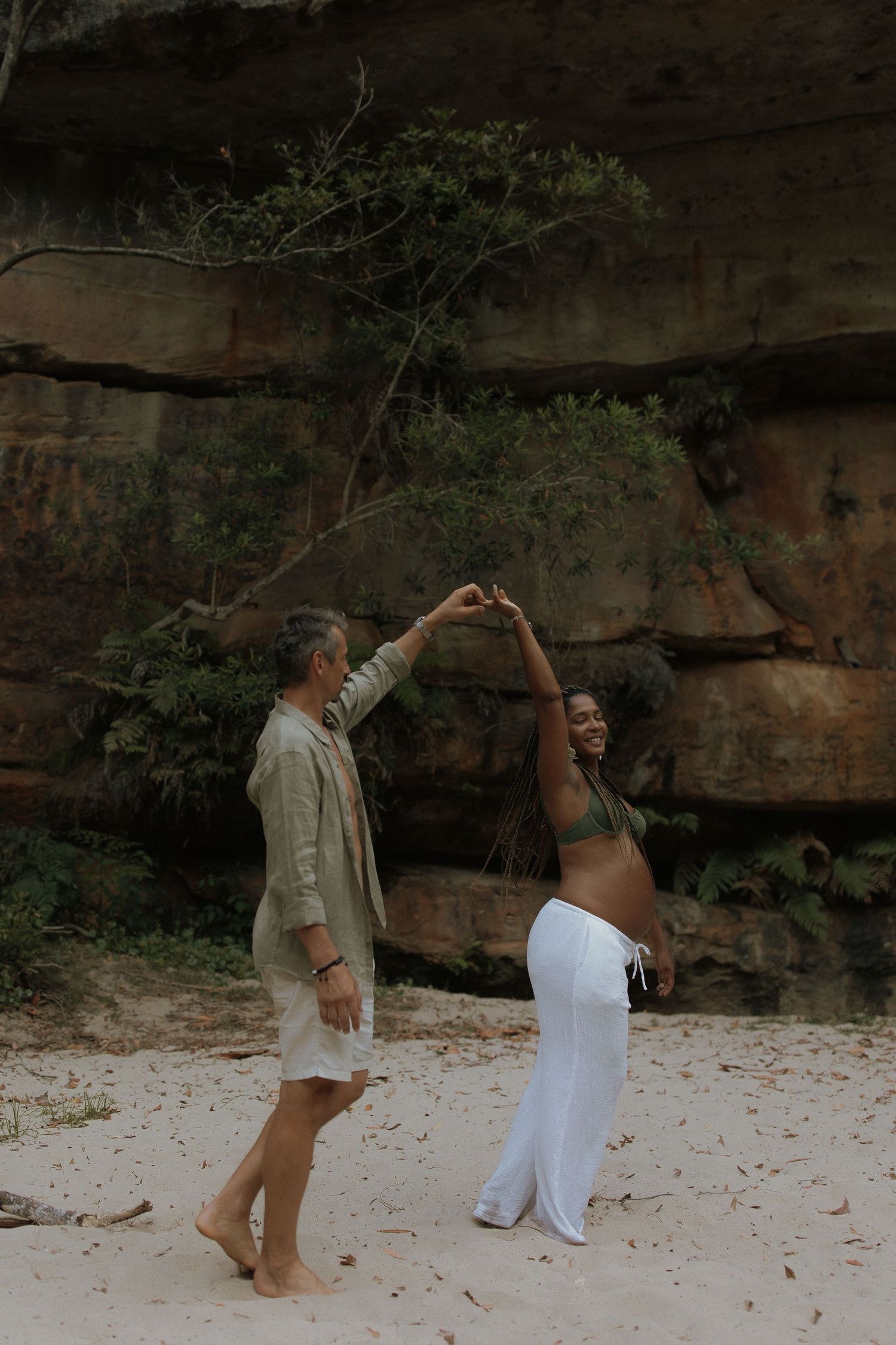 Two people dancing together on a sandy beach with rocky cliffs in the background.