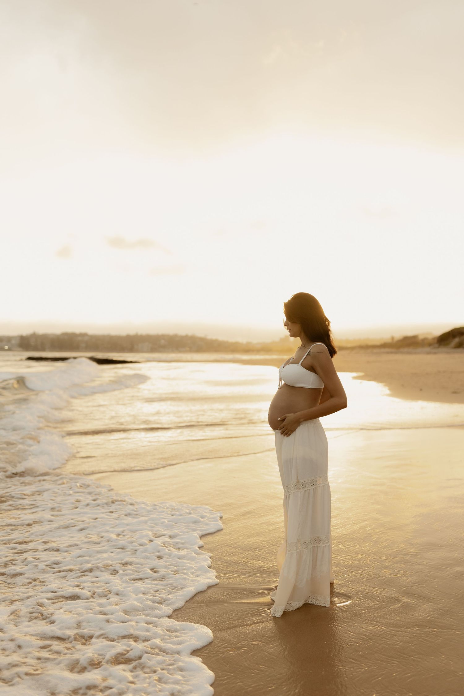 Silhouette of pregnant woman standing at ocean's edge during golden sunset.
