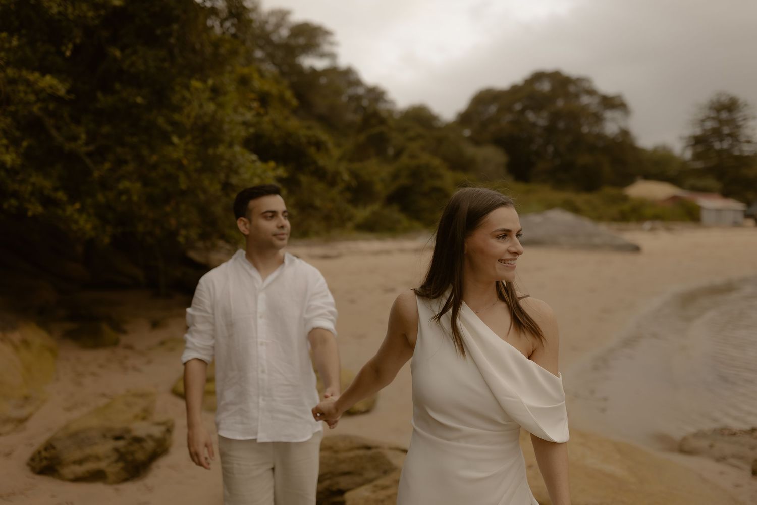 A couple walks hand in hand along a sandy beach with lush greenery behind them.