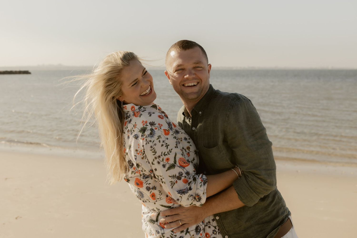 A couple embraces and laughs together while walking along a sandy beach at sunset.