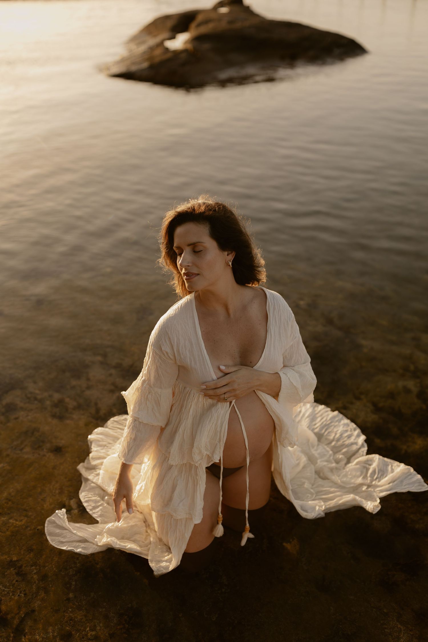 A figure in a flowing white dress kneels in shallow water during golden hour with rocks visible in background.