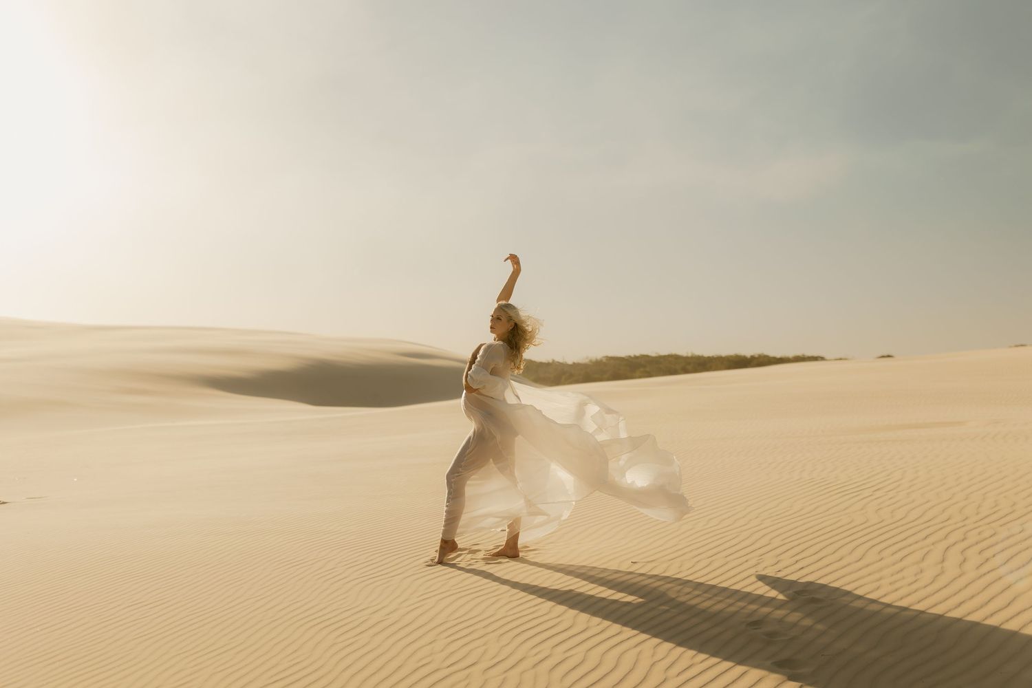 Woman in flowing white dress dancing gracefully on golden sand dunes under a soft, hazy sky.