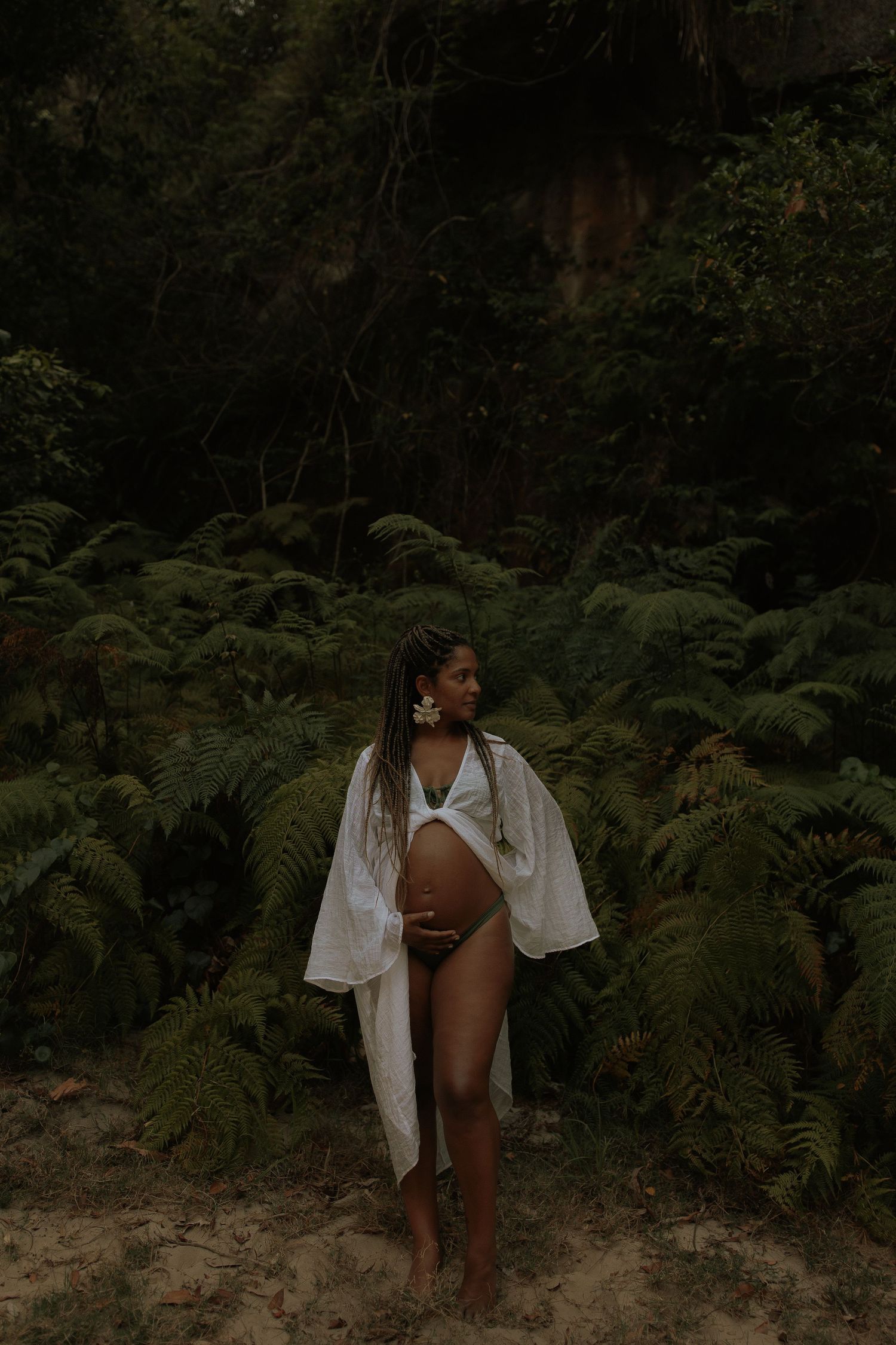 Pregnant woman in white robe and bikini posing among dense green ferns in a forest.