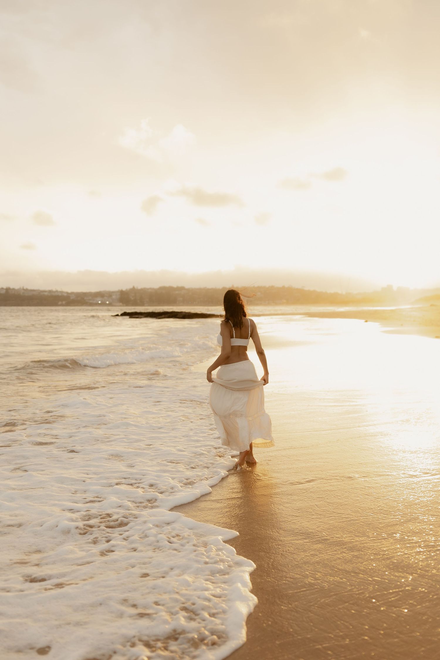 Pregnant woman in white crop top and skirt walks along shoreline as golden sunset waves roll in.
