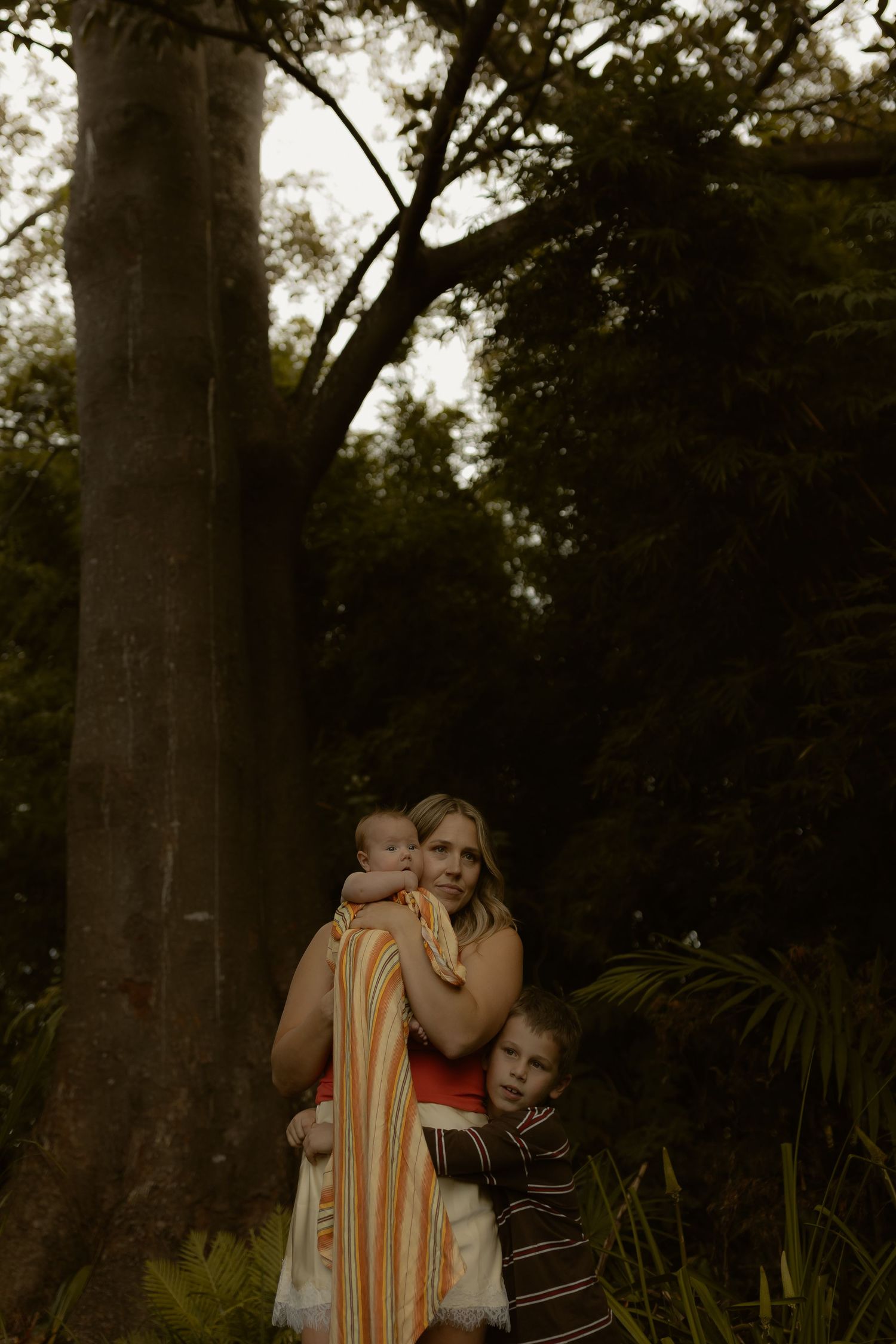 A smiling mother holding a baby in a striped wrap while a young child stands beside her in a forest.