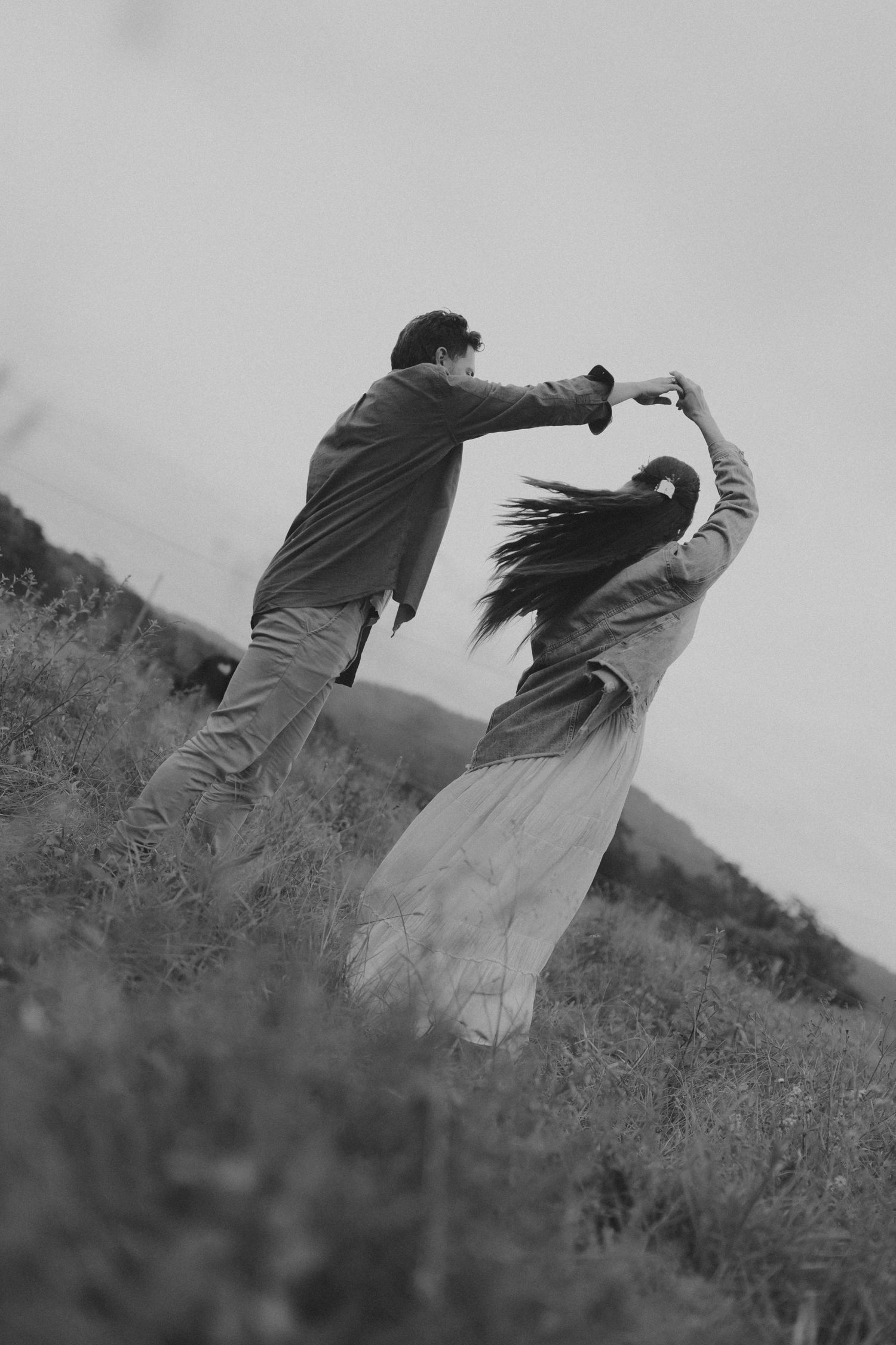 Black and white photo of a couple dancing in a field, man spinning woman in a flowing dress outdoors.