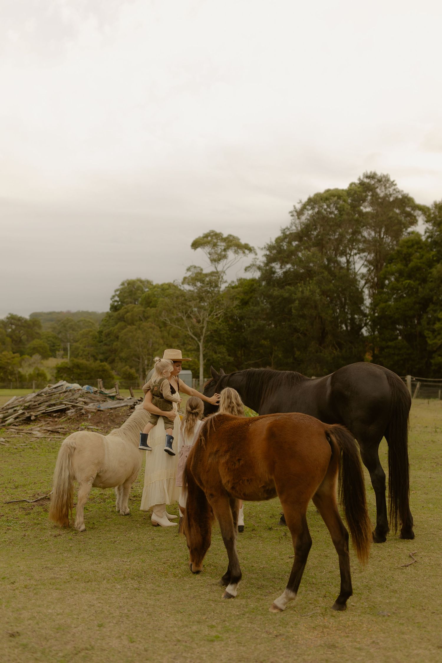 A woman in a cream dress and hat stands among brown and black horses with a child on a grassy farm.