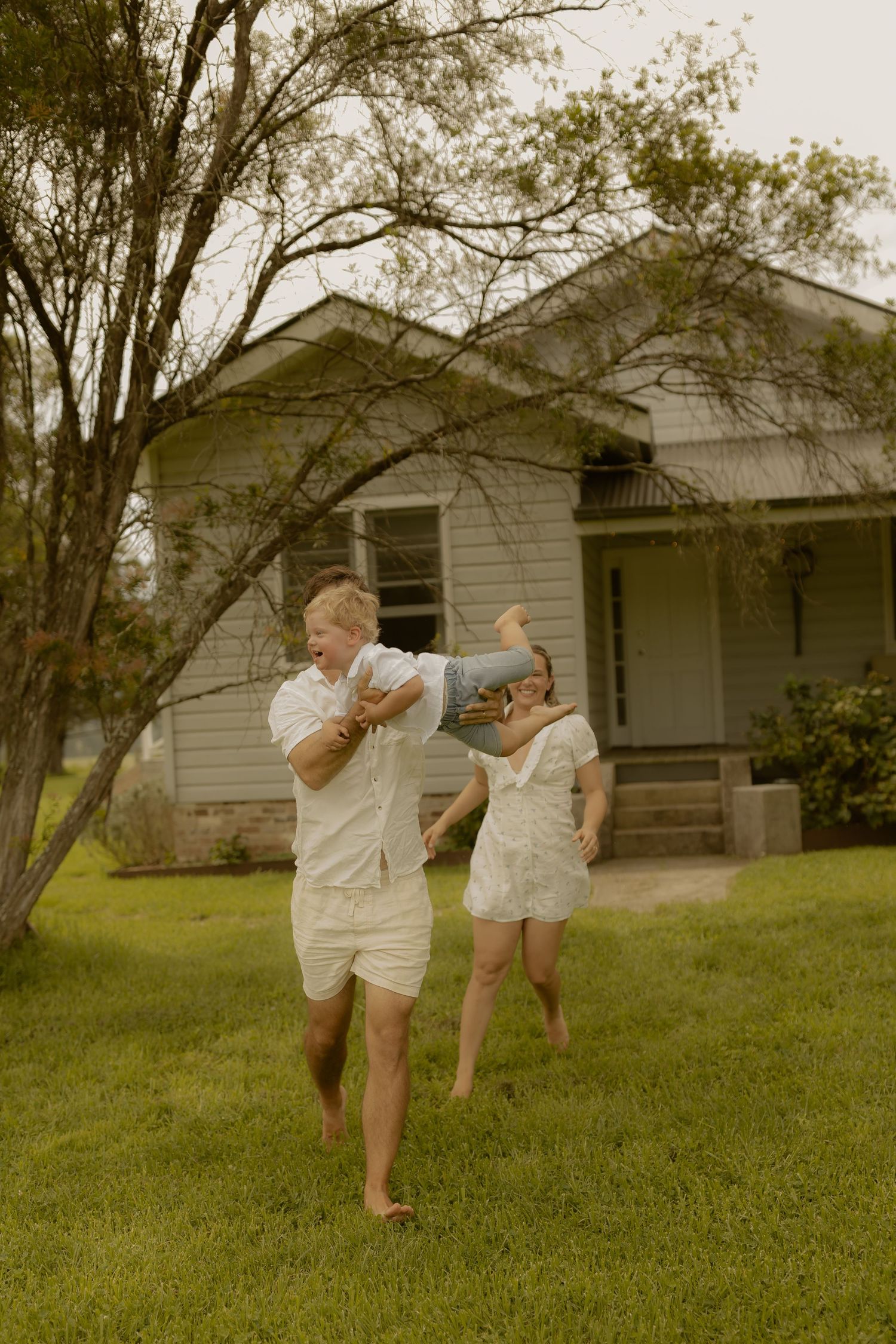 Two people in white outfits dance playfully on a lawn in front of a rustic house.