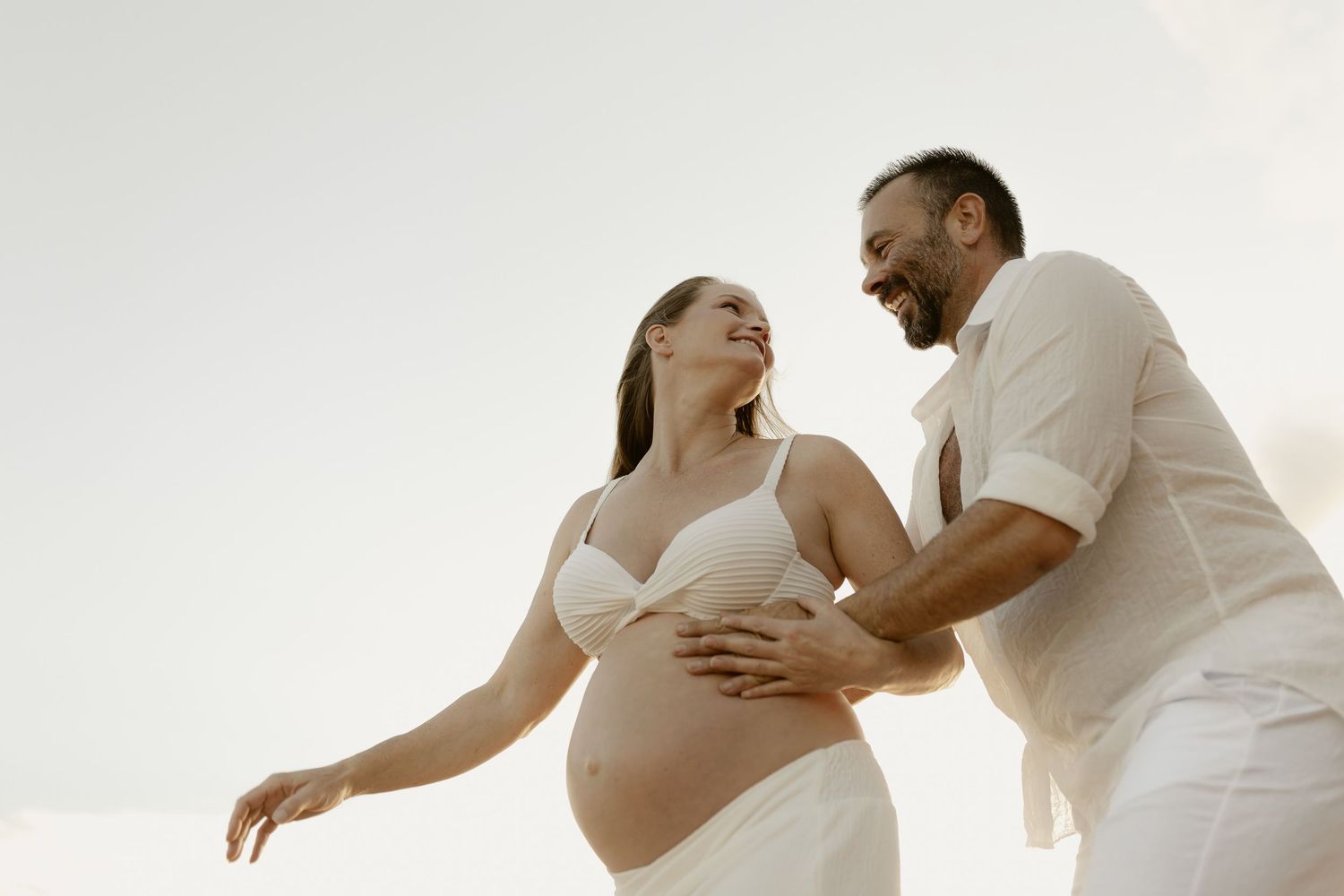 Romantic couple in white clothing embraces while dancing together in an elegant pose against light background.