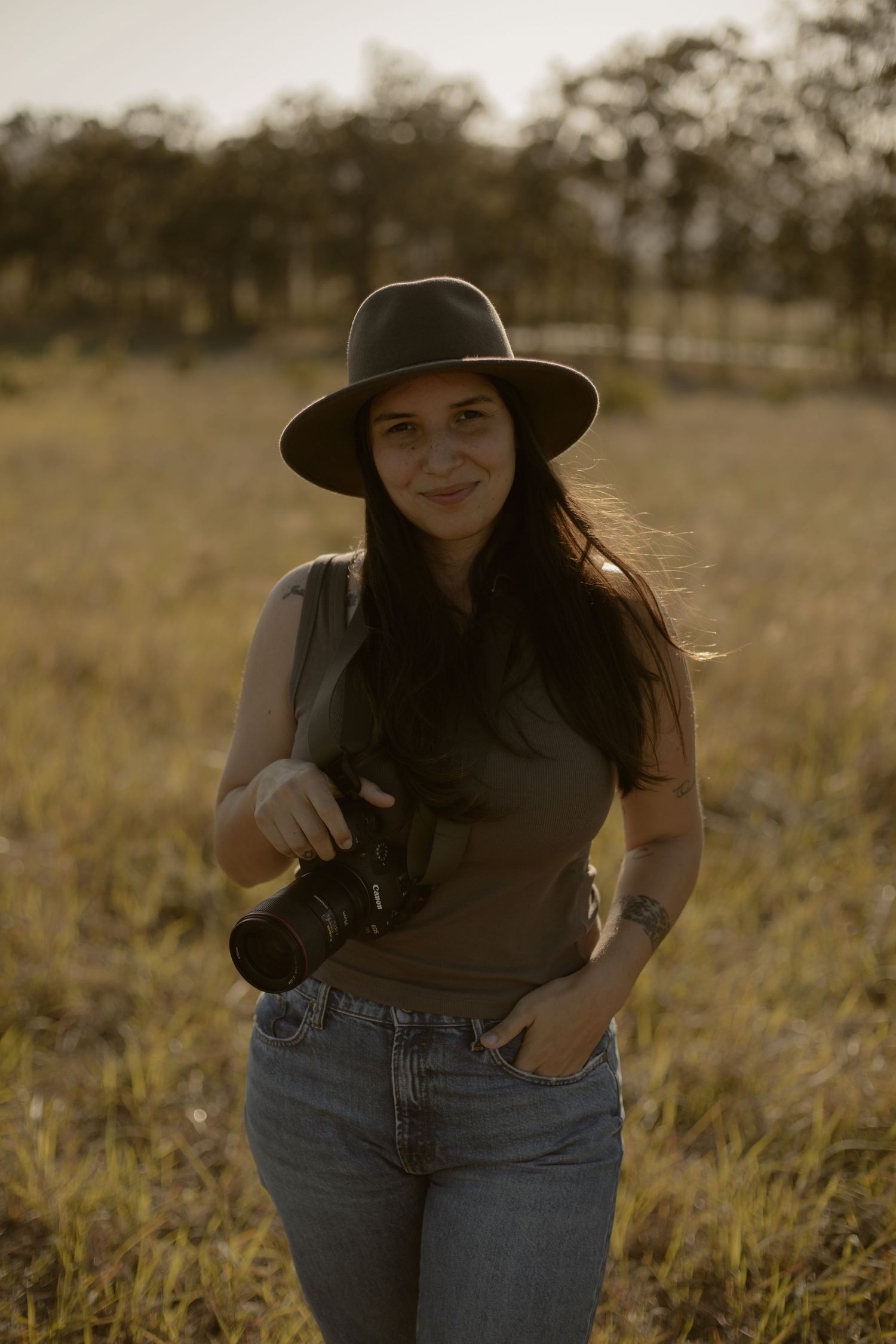Person wearing a brown hat and dark top poses in a sunlit field with tall grass at golden hour.