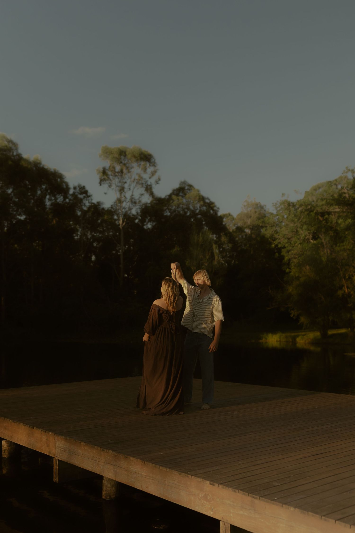 Couple standing on a wooden deck at sunset, surrounded by lush green trees in warm evening light.