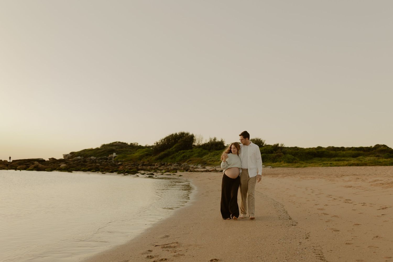 Expecting couple walking hand-in-hand on sandy beach at sunset near rocky shoreline.