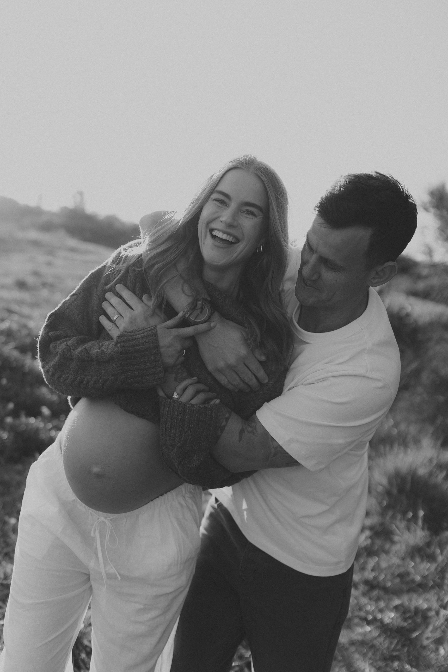 Black and white photo of a laughing pregnant woman being embraced by a man outdoors in a field.