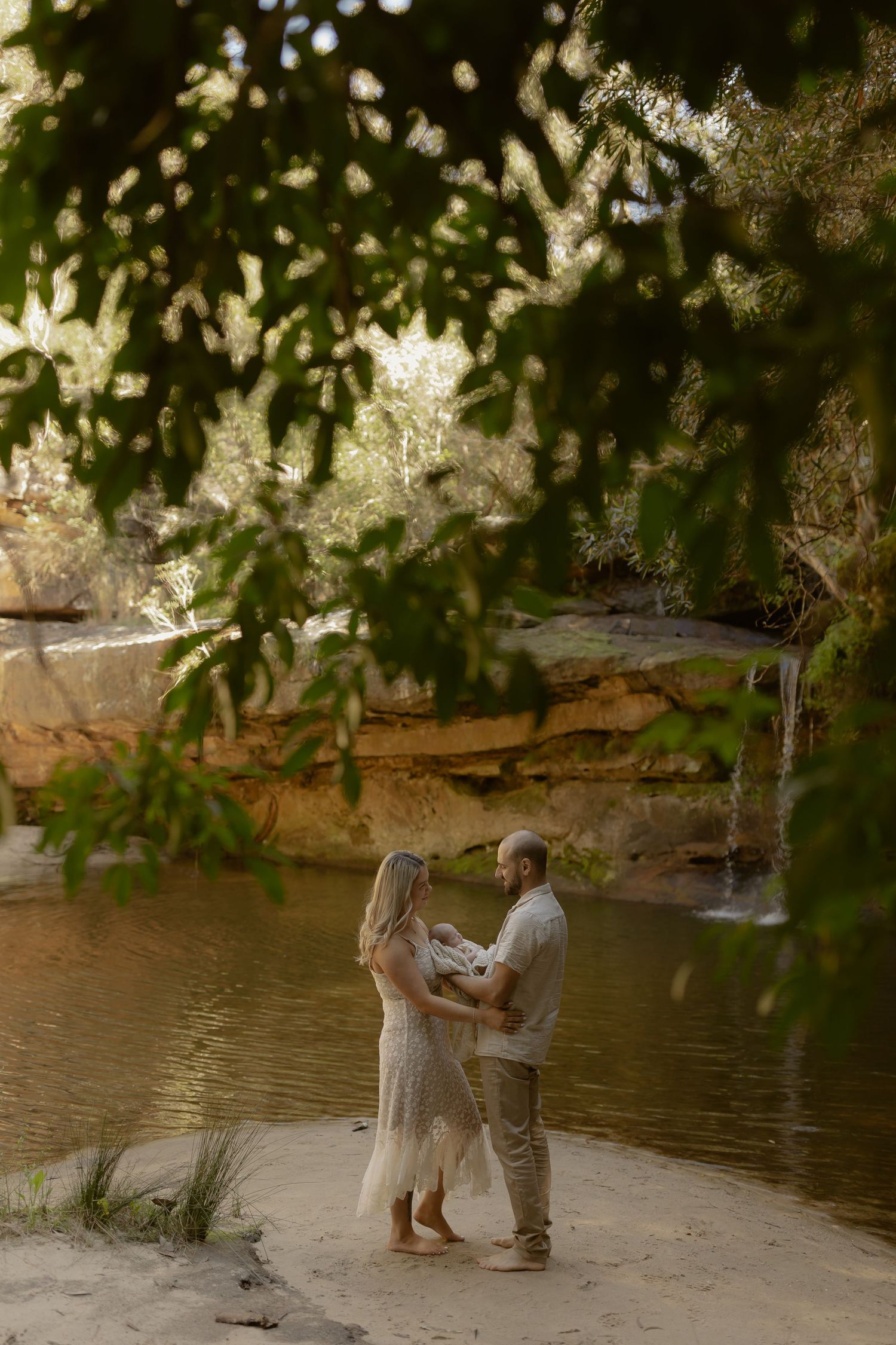 Couple standing barefoot by a serene river surrounded by lush greenery, sharing a tender moment during an outdoor photoshoot.