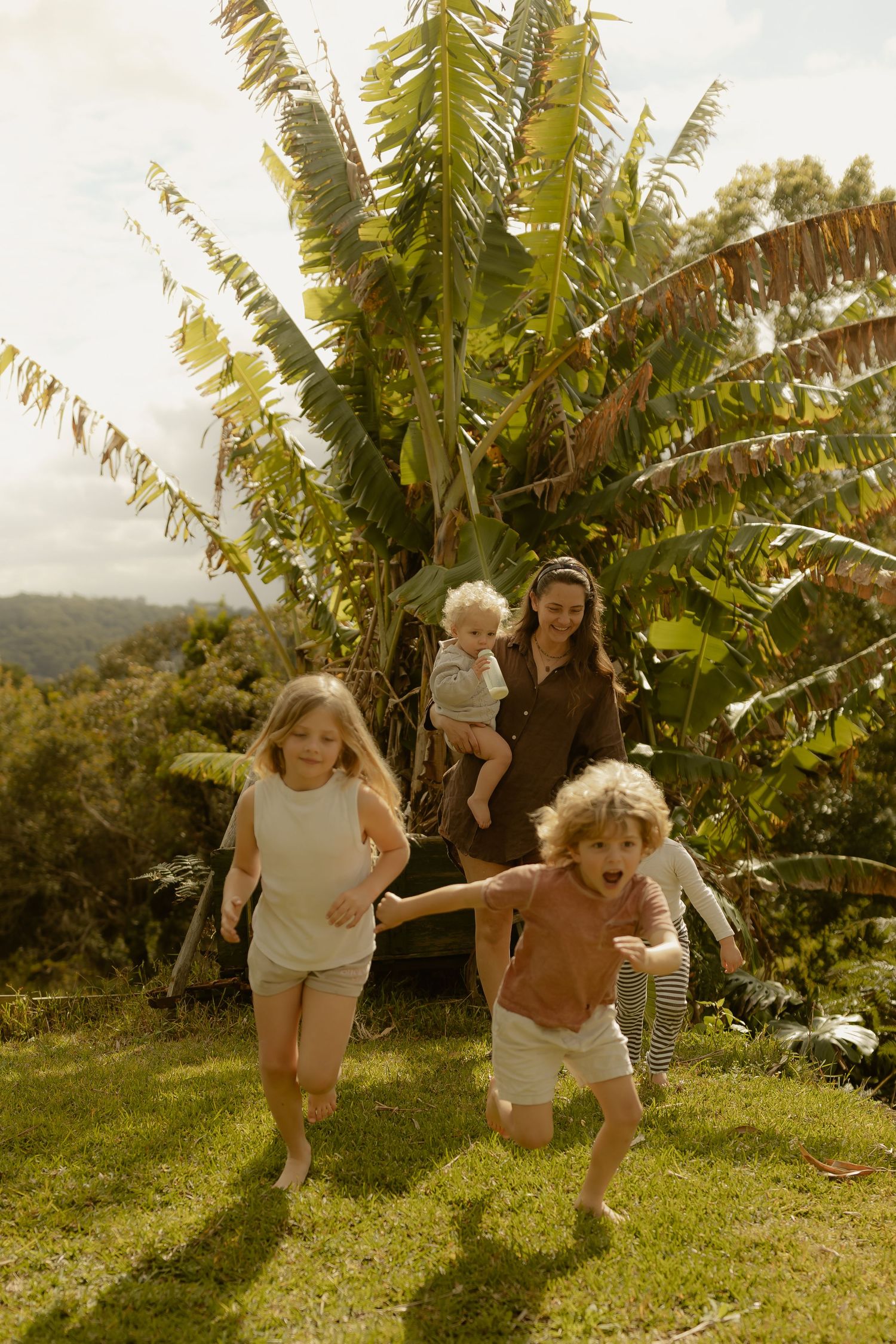 Mother holding baby watches two children run across green lawn with tropical trees behind.