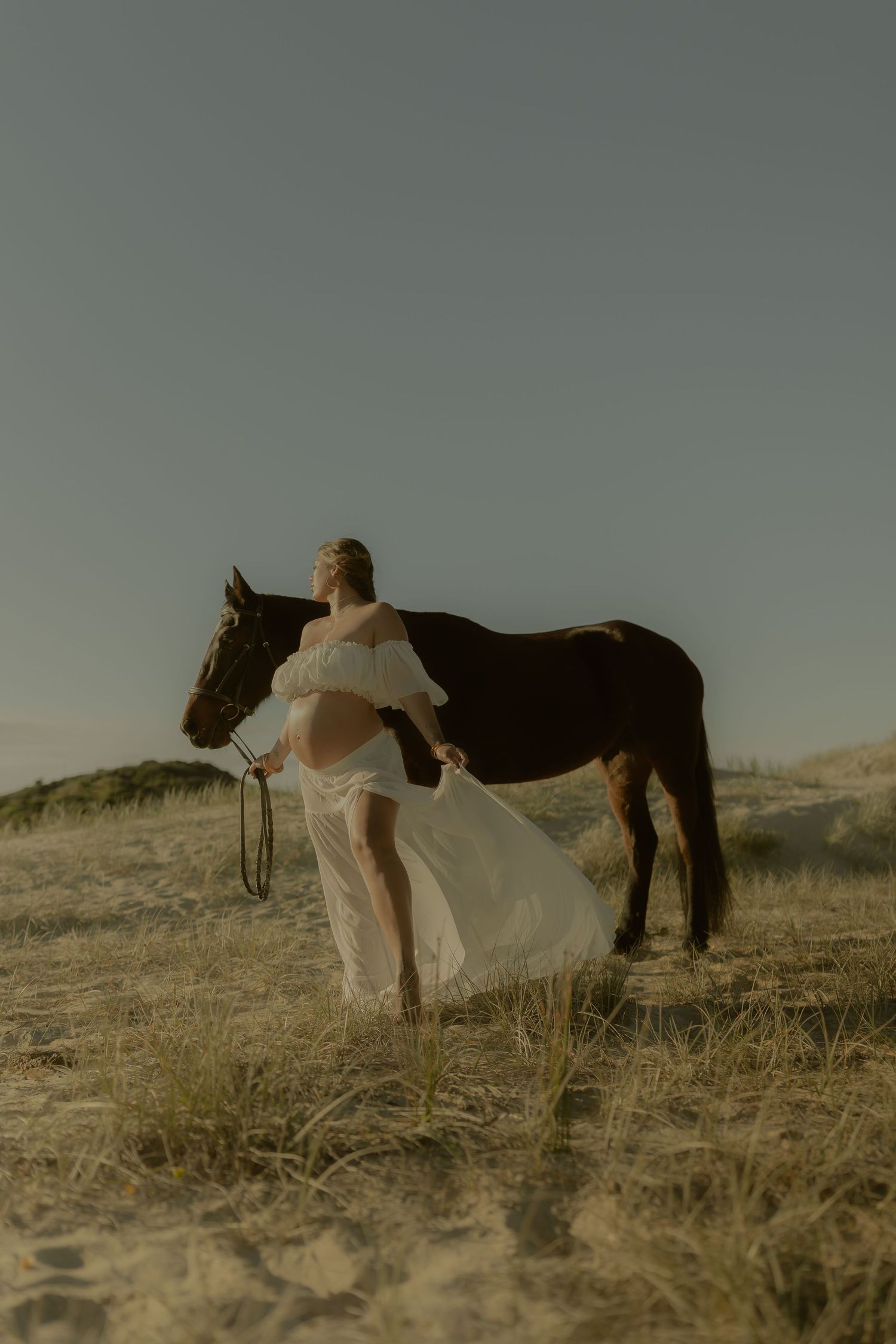 Pregnant woman in flowing white dress stands beside a dark horse on sandy dunes under a clear sky.