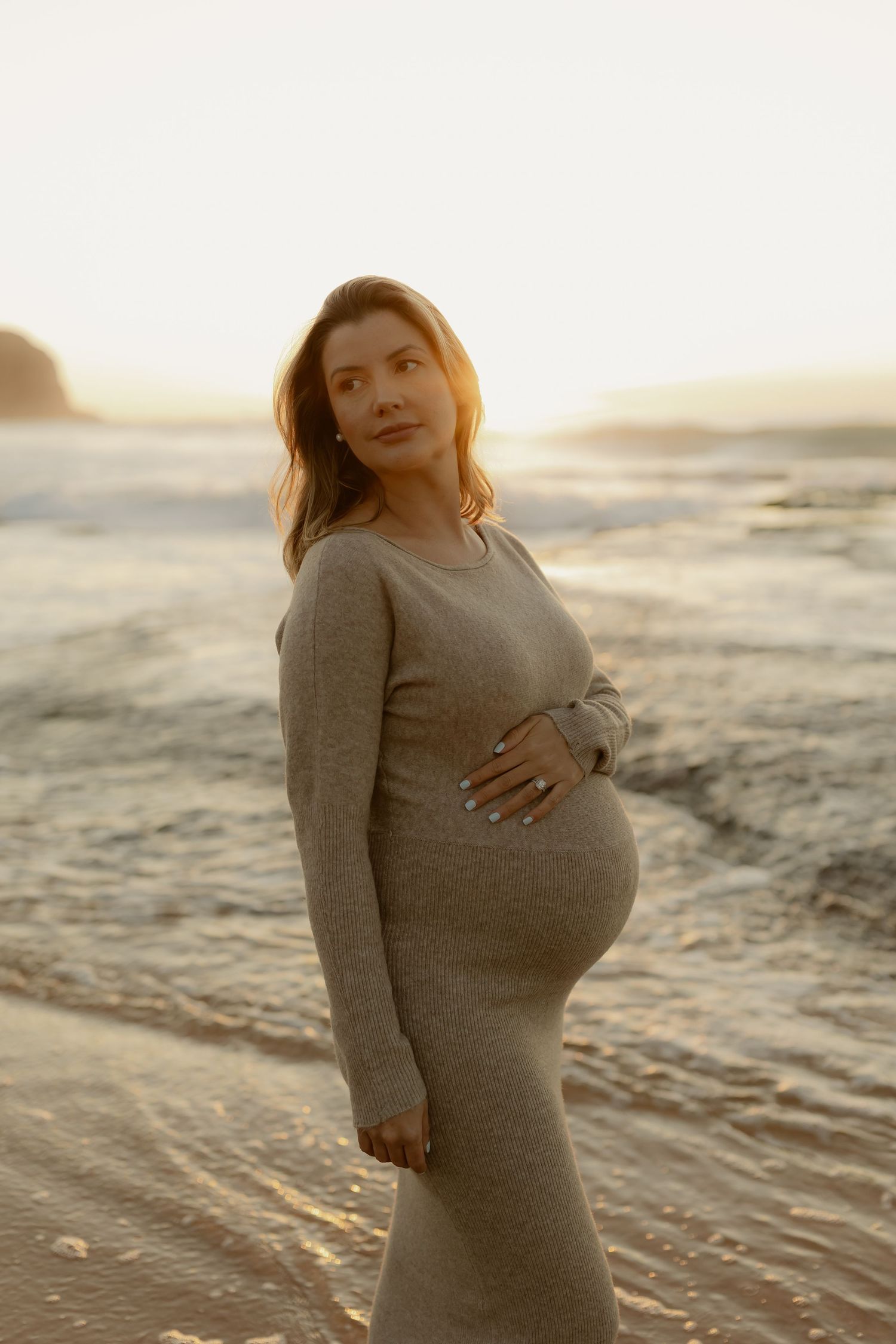 Pregnant woman in a gray knit dress cradling her baby bump on a beach at golden sunset.