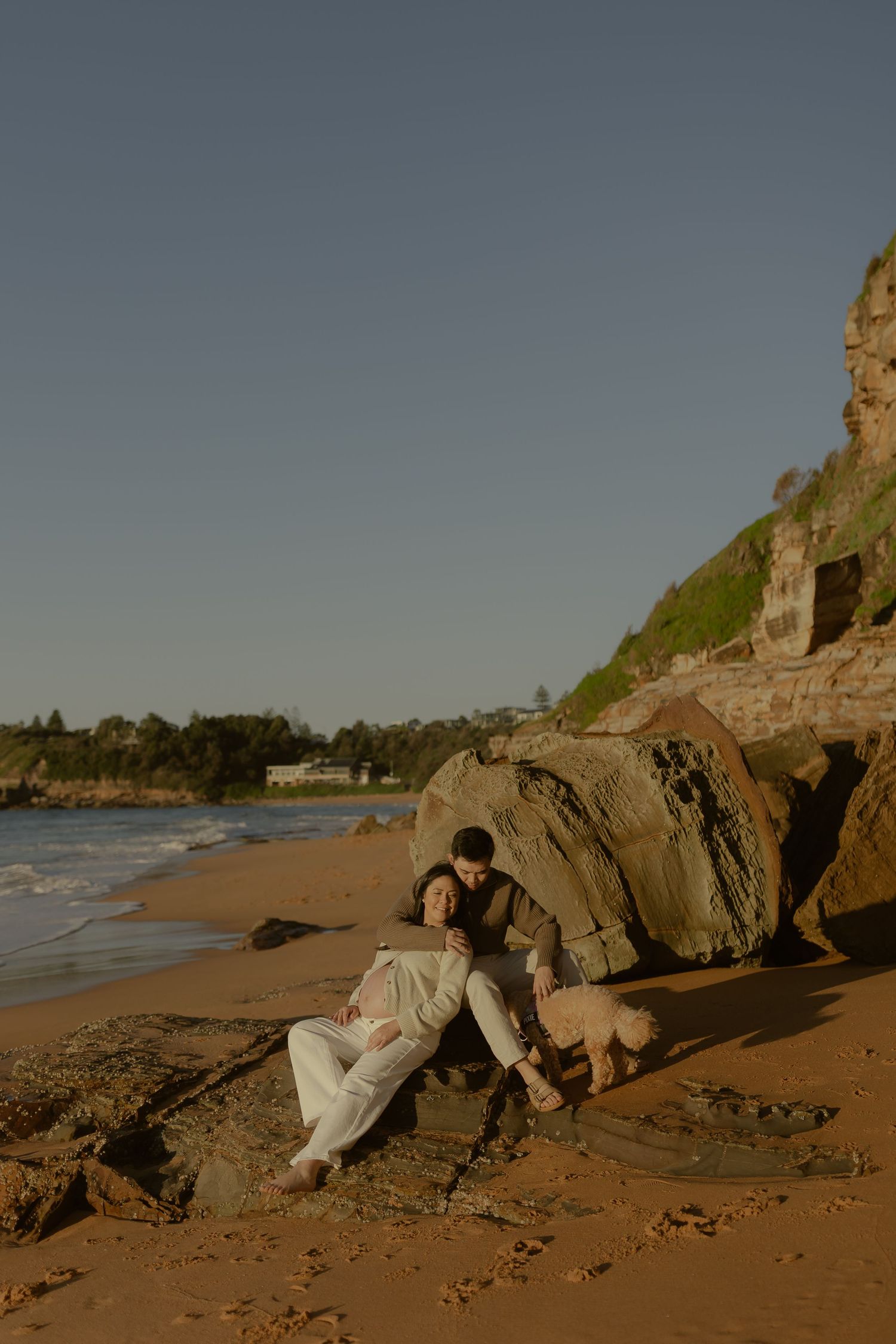 Couple relaxing on rocky beach at golden hour, woman leaning back on man near large boulders with ocean waves behind.