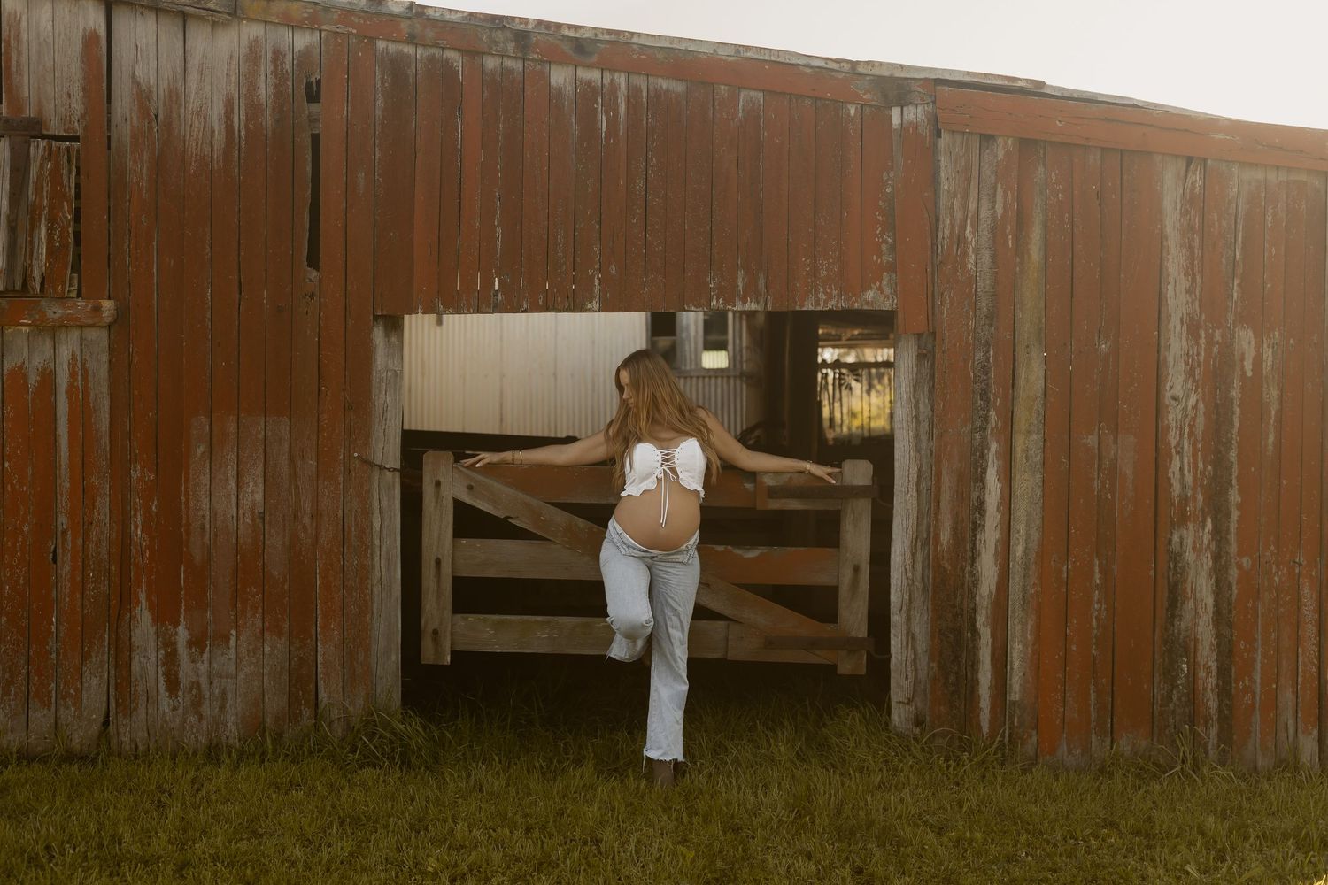 Pregnant woman in white top and jeans standing in rustic red barn doorway on green grass.