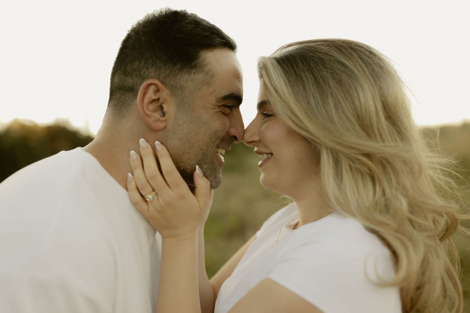 Romantic couple sharing a tender moment outdoors, almost kissing, woman wearing engagement ring, soft natural lighting.