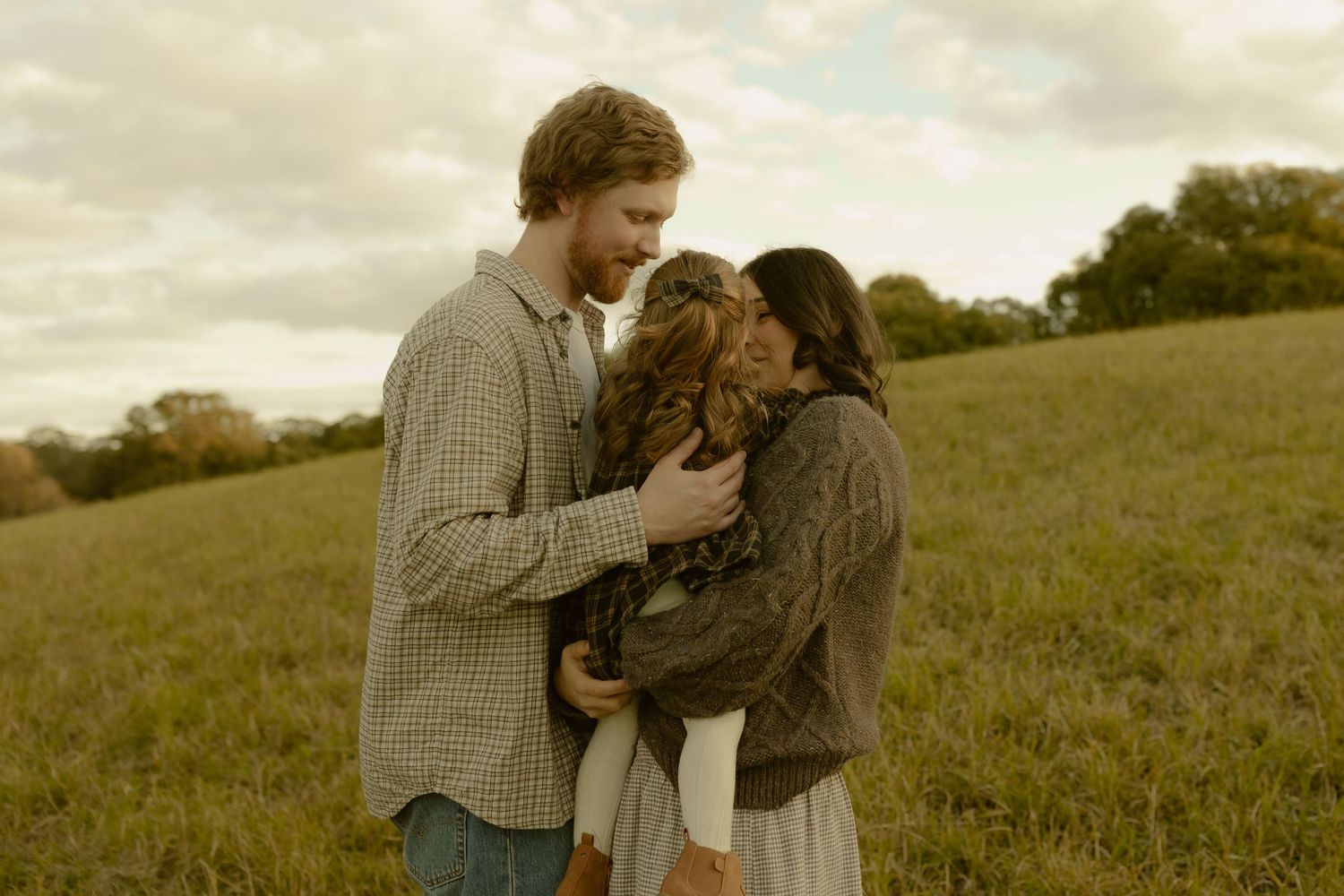 A couple shares a tender moment embracing a child in a green field under a cloudy sky.