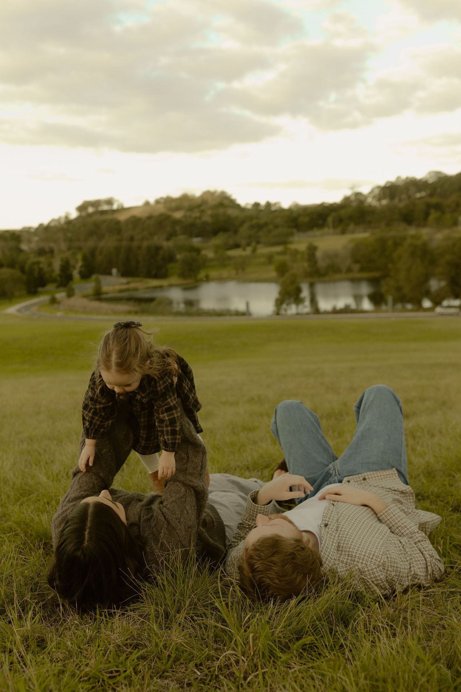 Family relaxing on green grass near a lake, with a child playing over two adults lying down in a scenic park.