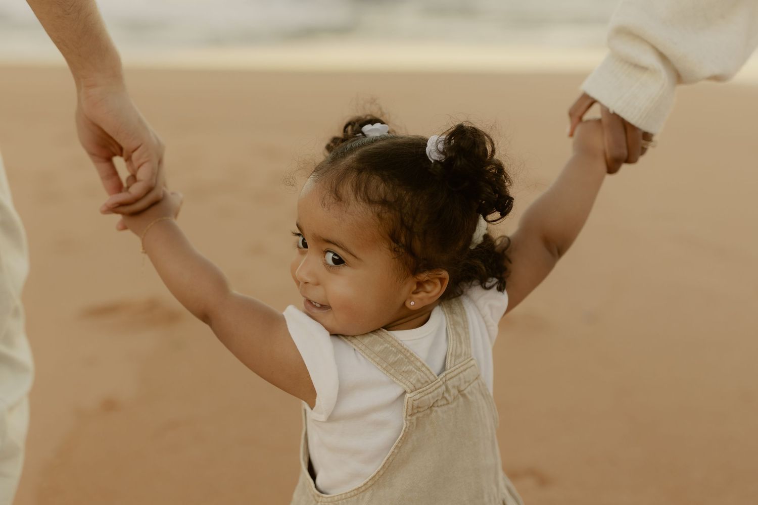 A toddler girl in overalls holds both parents hands on a sandy beach, looking up with a joyful smile.
