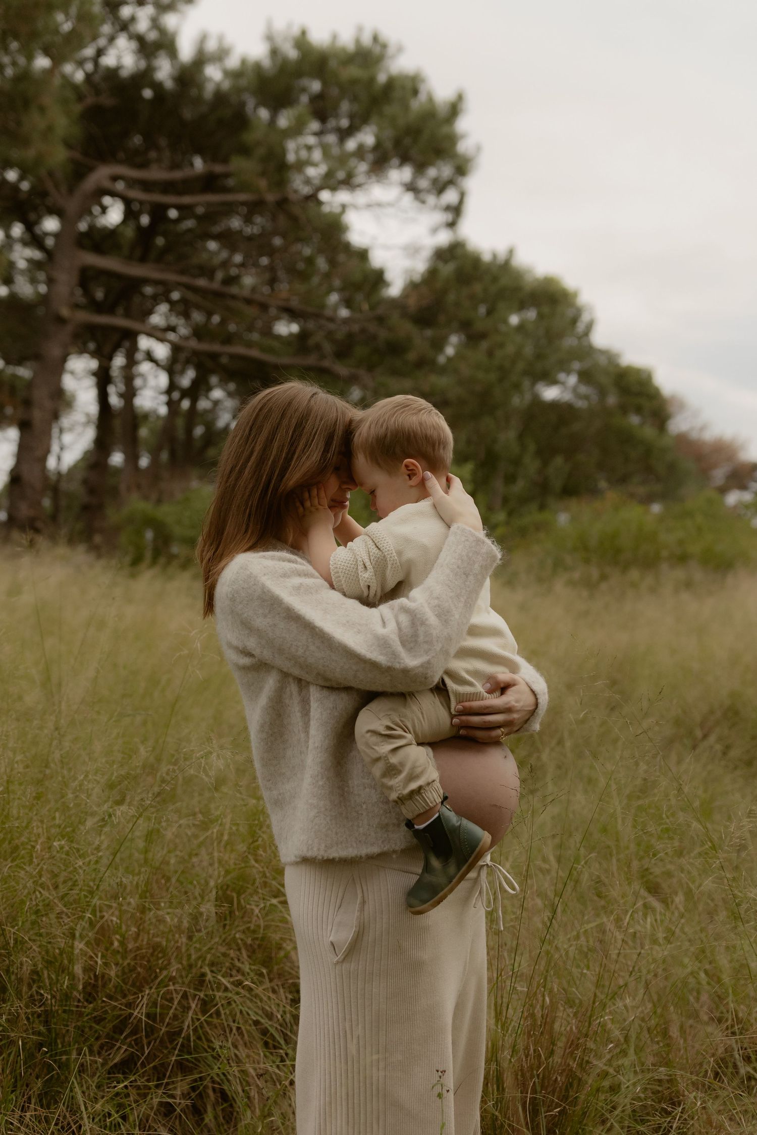 Pregnant woman in neutral outfit holding toddler in green field with trees, tender mother and child moment outdoors.
