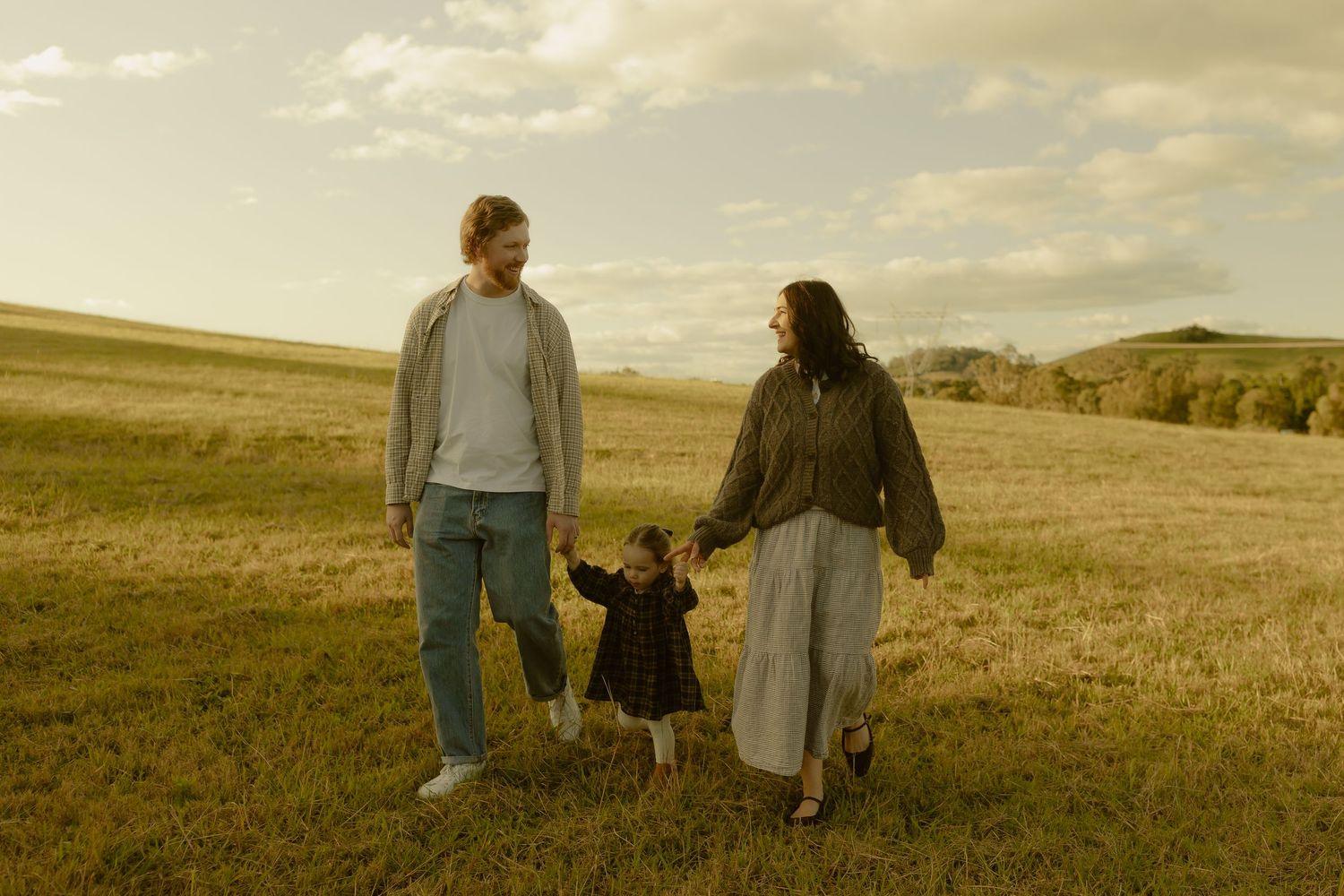 A young family holds hands in a green field at golden hour, parents smiling at their toddler child.
