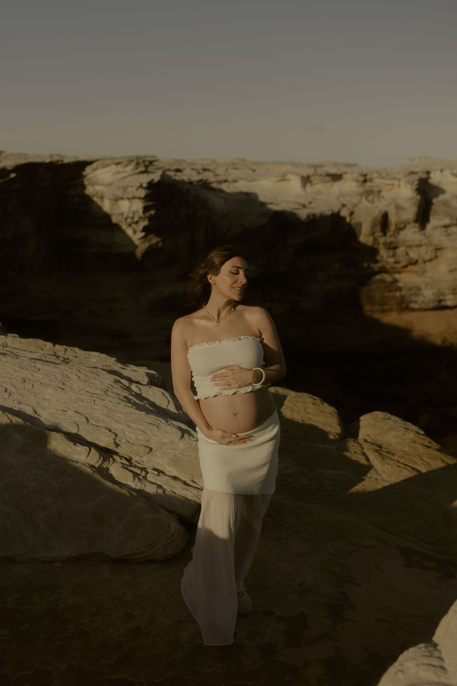 Pregnant woman in white crop top and skirt cradling her belly among sunlit desert rock formations.