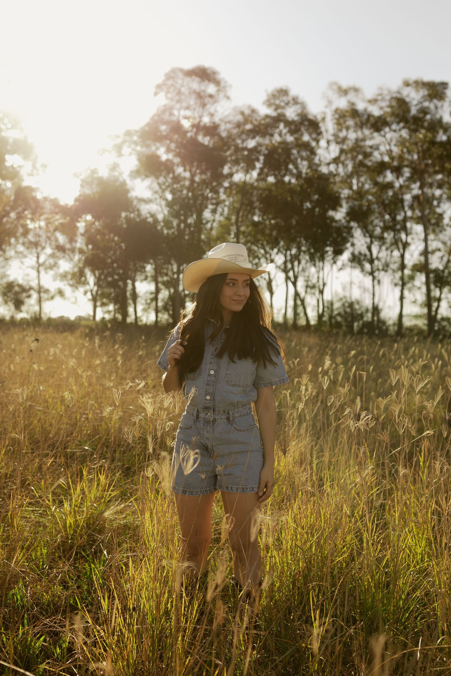 Young woman in denim romper and straw cowboy hat standing in golden sunlit field with trees in background.