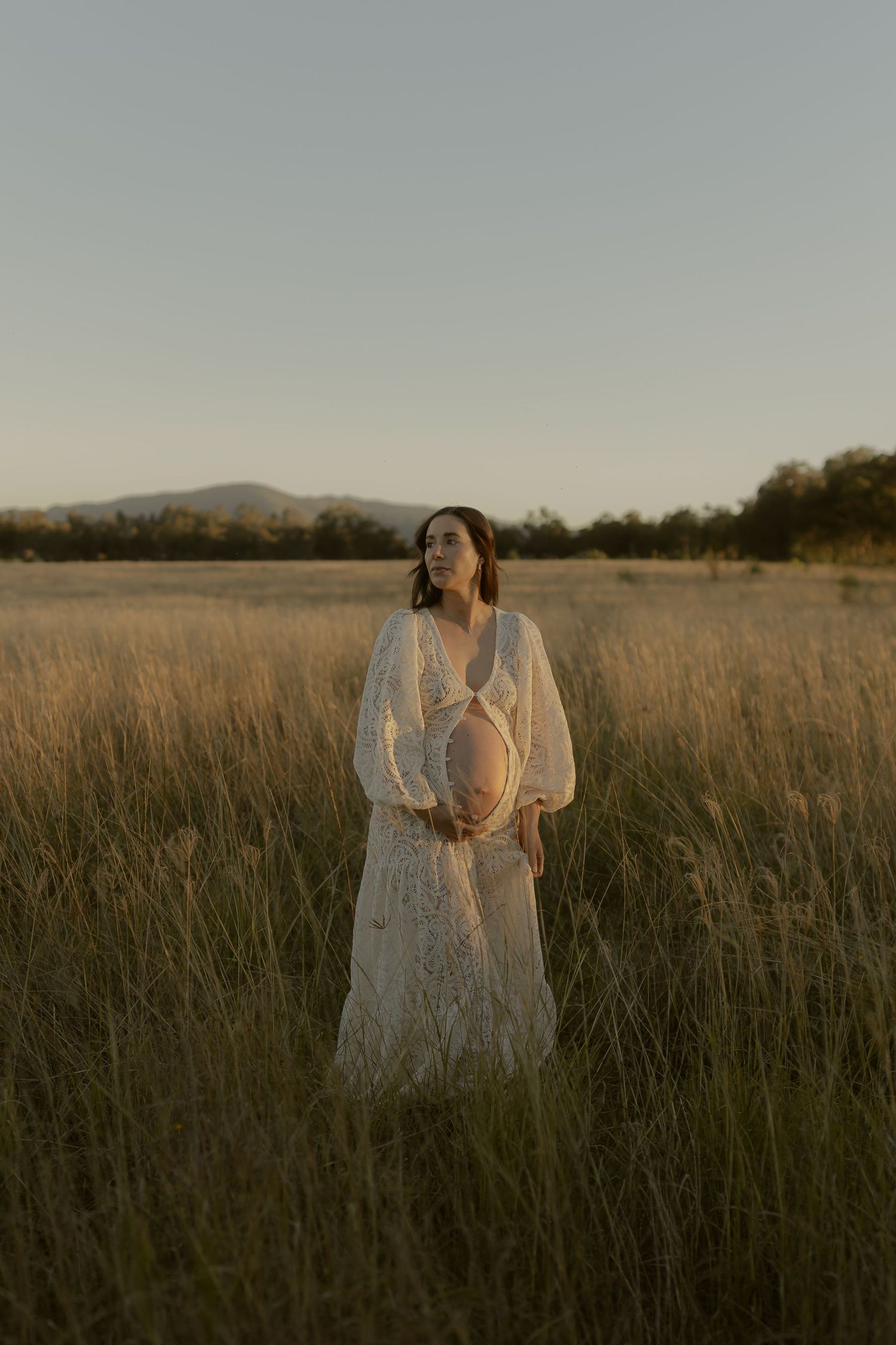 Pregnant woman in white lace dress standing in tall golden grass field at dusk.