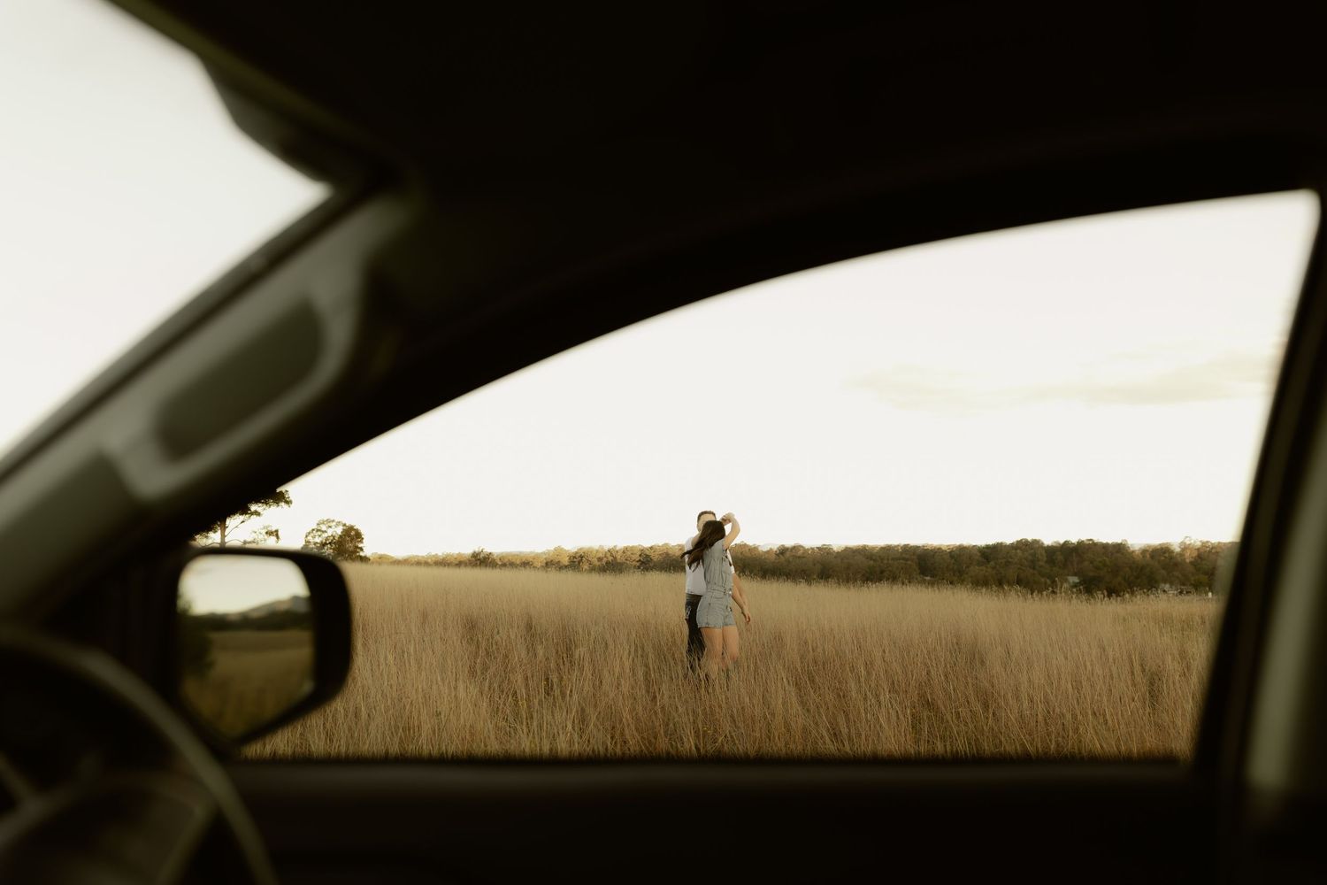 Woman standing alone in golden field at dusk, photographed through a car window frame.