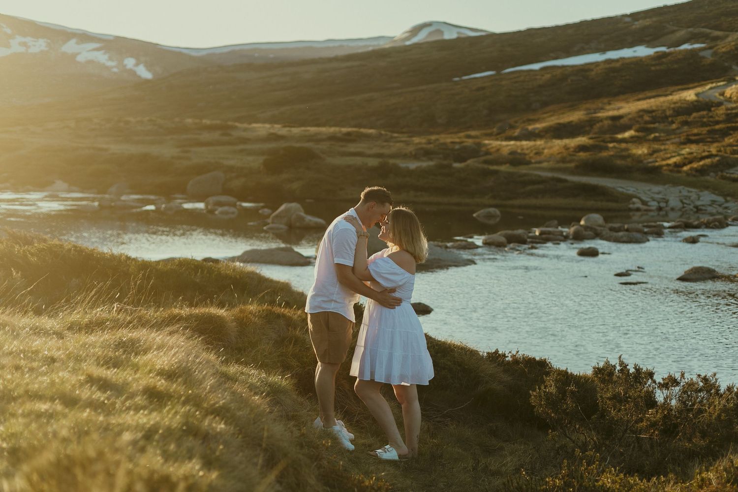A couple embraces on a scenic hillside overlooking a serene lake during golden hour.
