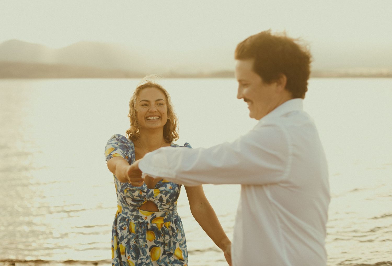 A couple dances together on a beach at sunset with soft golden light illuminating their joyful movements.