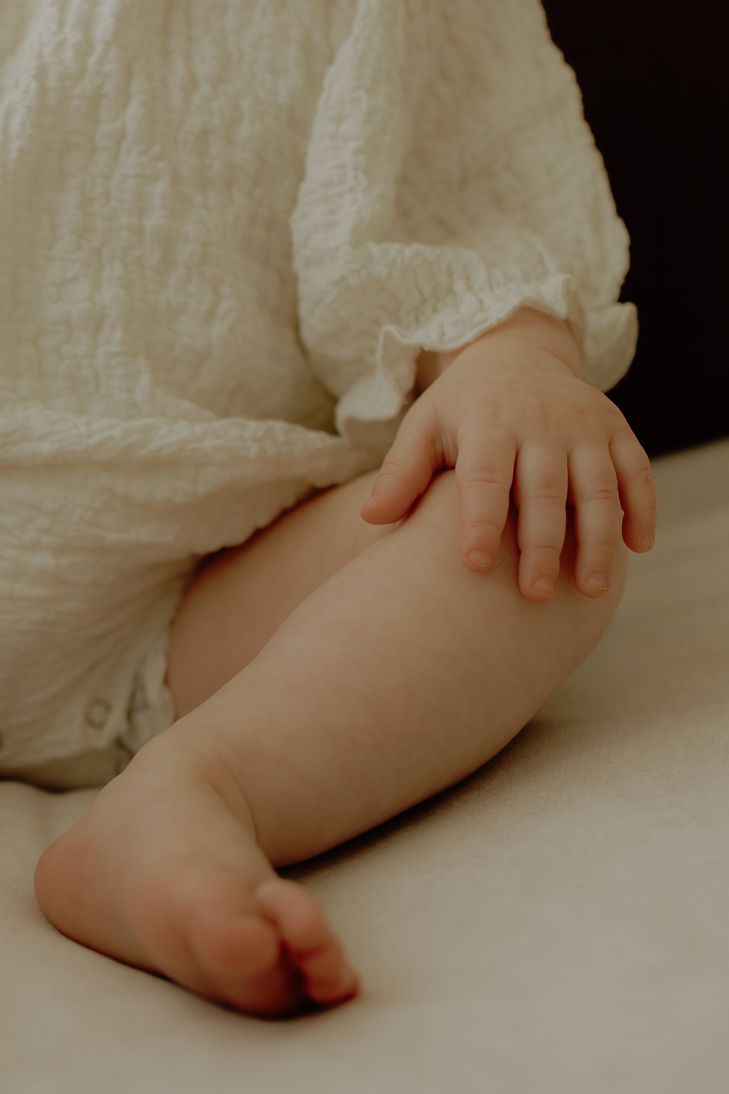 Close-up of a baby's chubby hand and bare leg resting on a soft surface, dressed in a white linen romper.