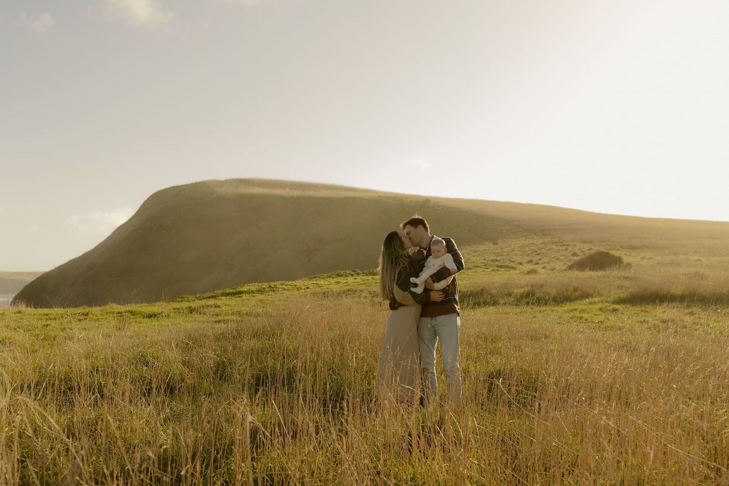 Couple embracing in a golden sunlit field with a green hillside backdrop.