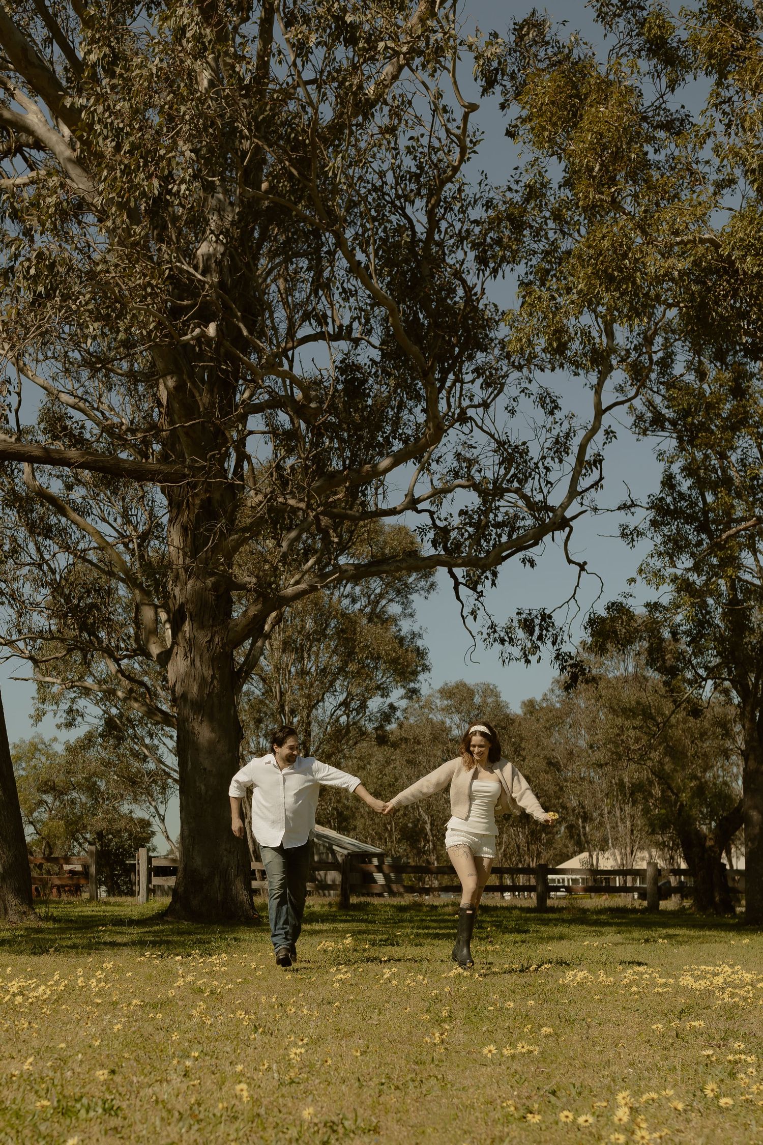 Two people playfully run and dance together across a grassy field with eucalyptus trees.