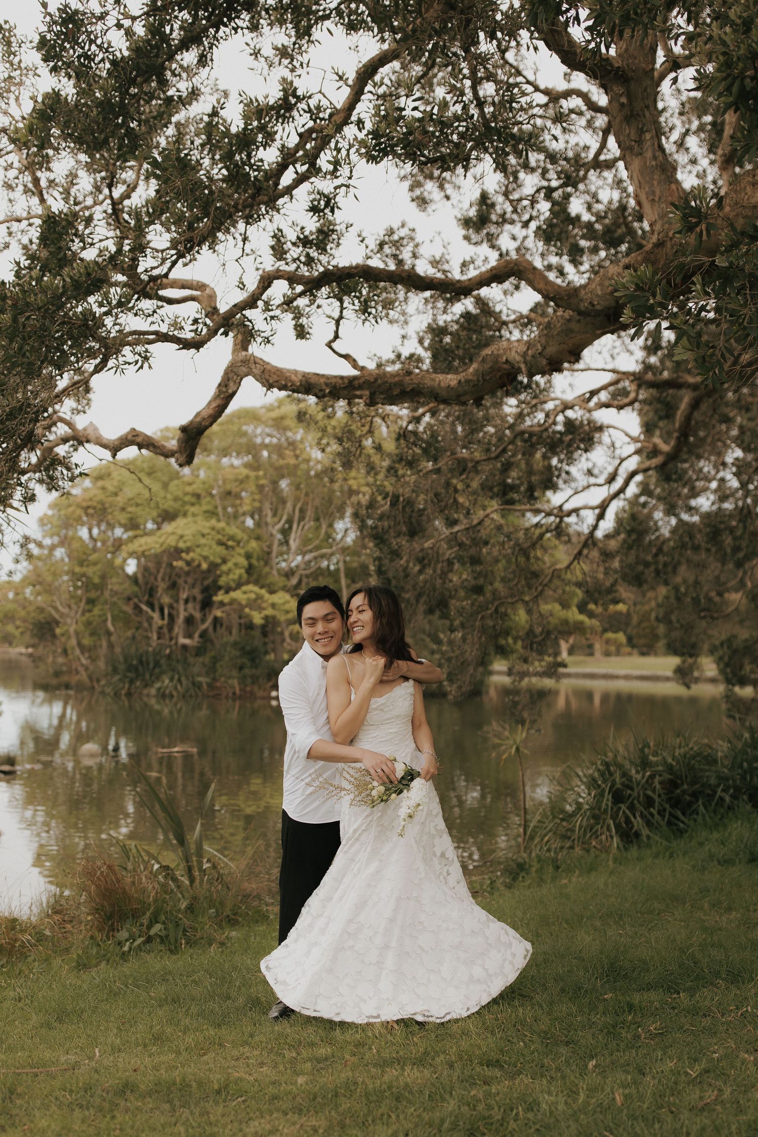 Romantic couple in white attire dancing and embracing under large oak trees near a serene lake.
