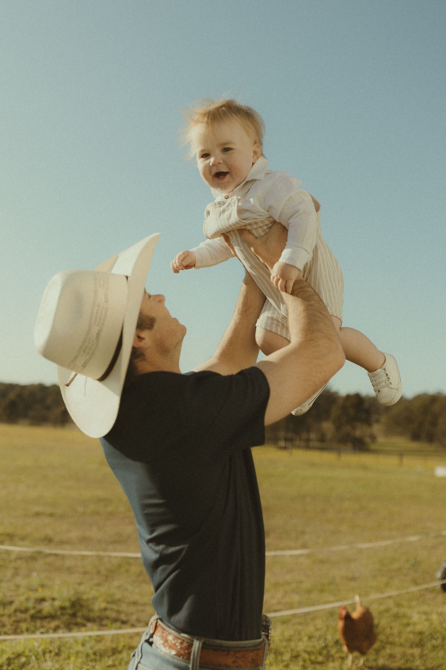 A playful moment in a sunny field as a baby is lifted high into the air while wearing a white hat.
