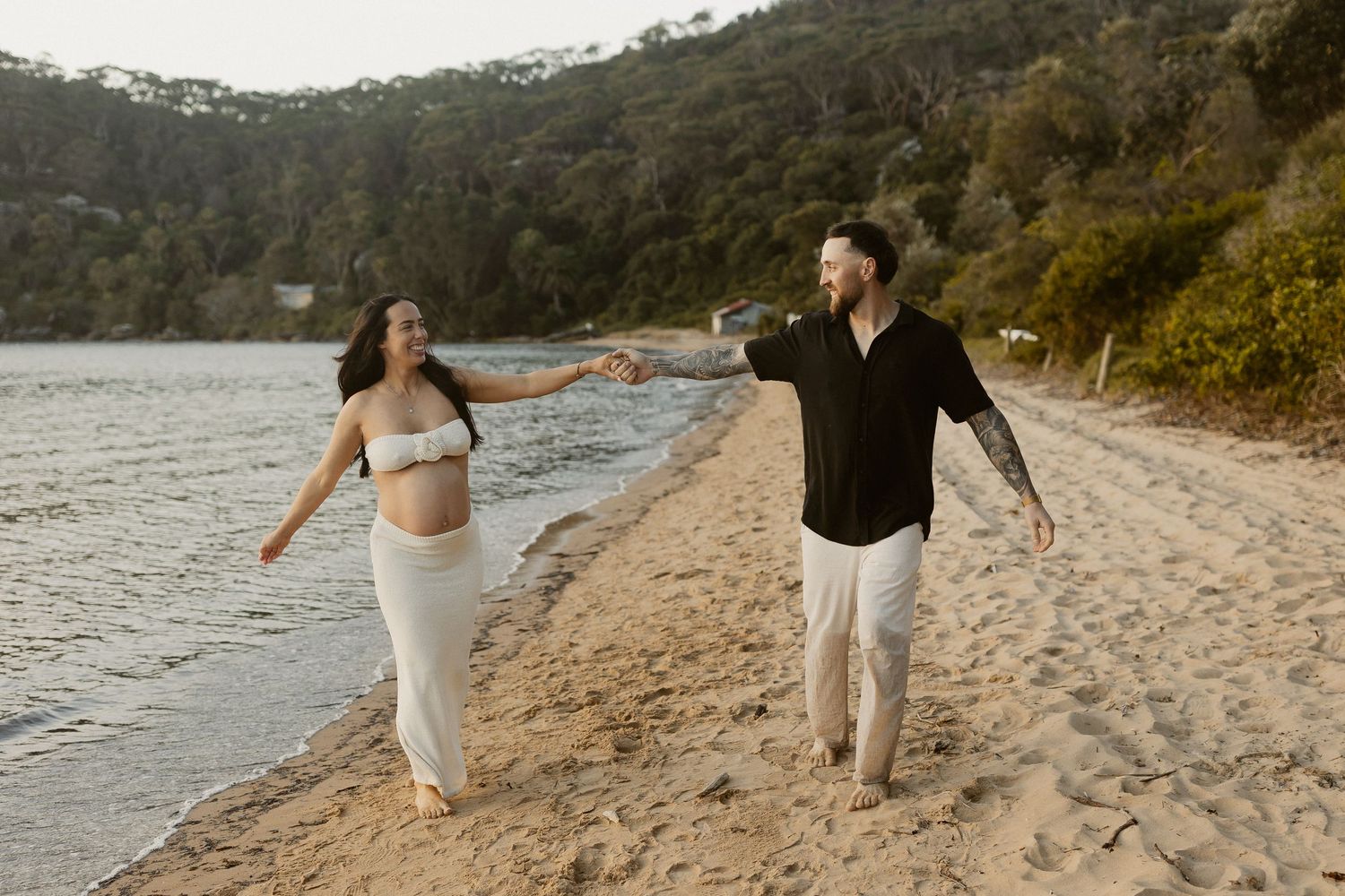 Couple walking hand in hand along a scenic beach with hills in the background.