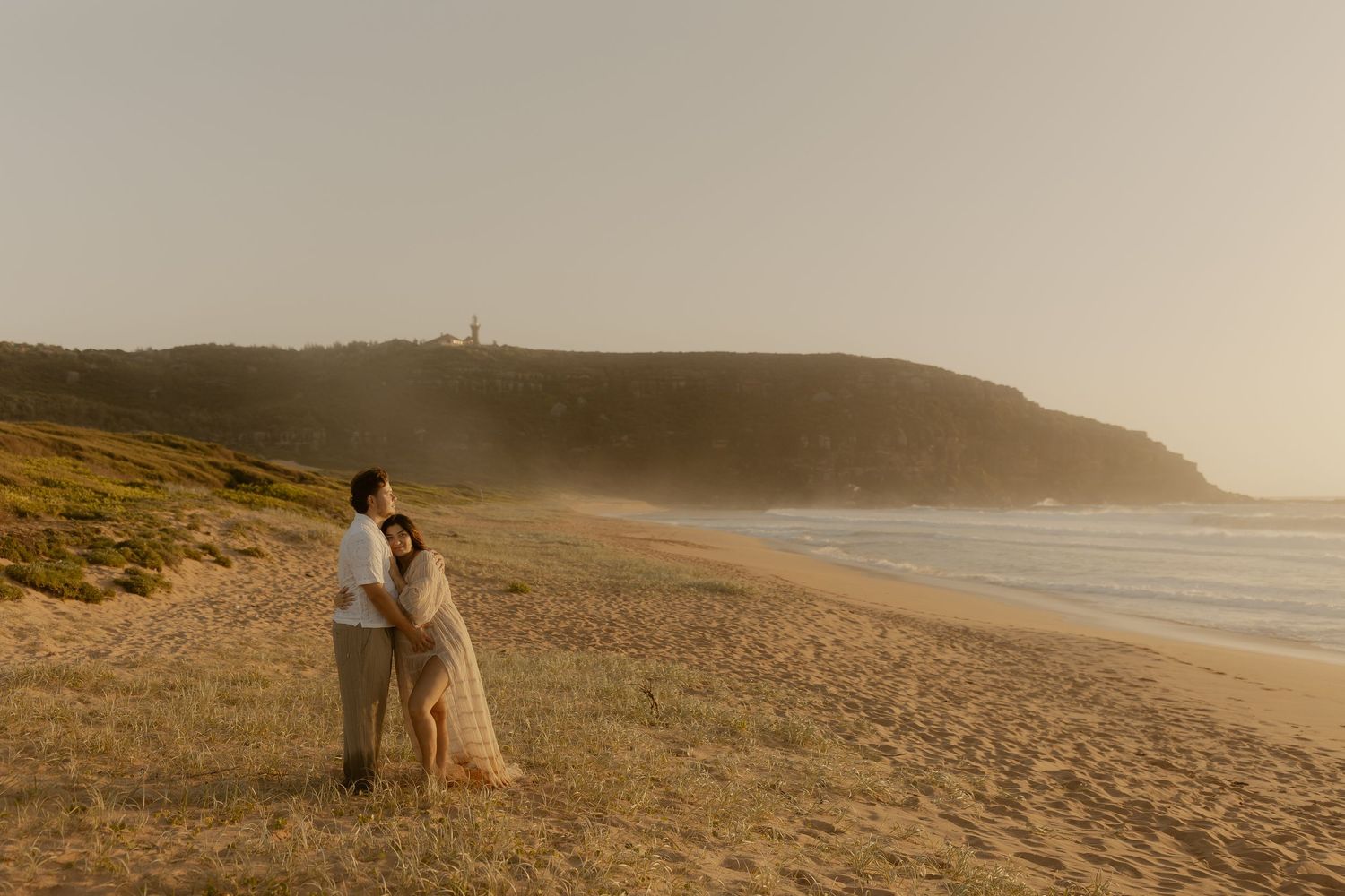 Romantic couple embraces on a misty coastal hillside at sunset with ocean waves crashing in the background.