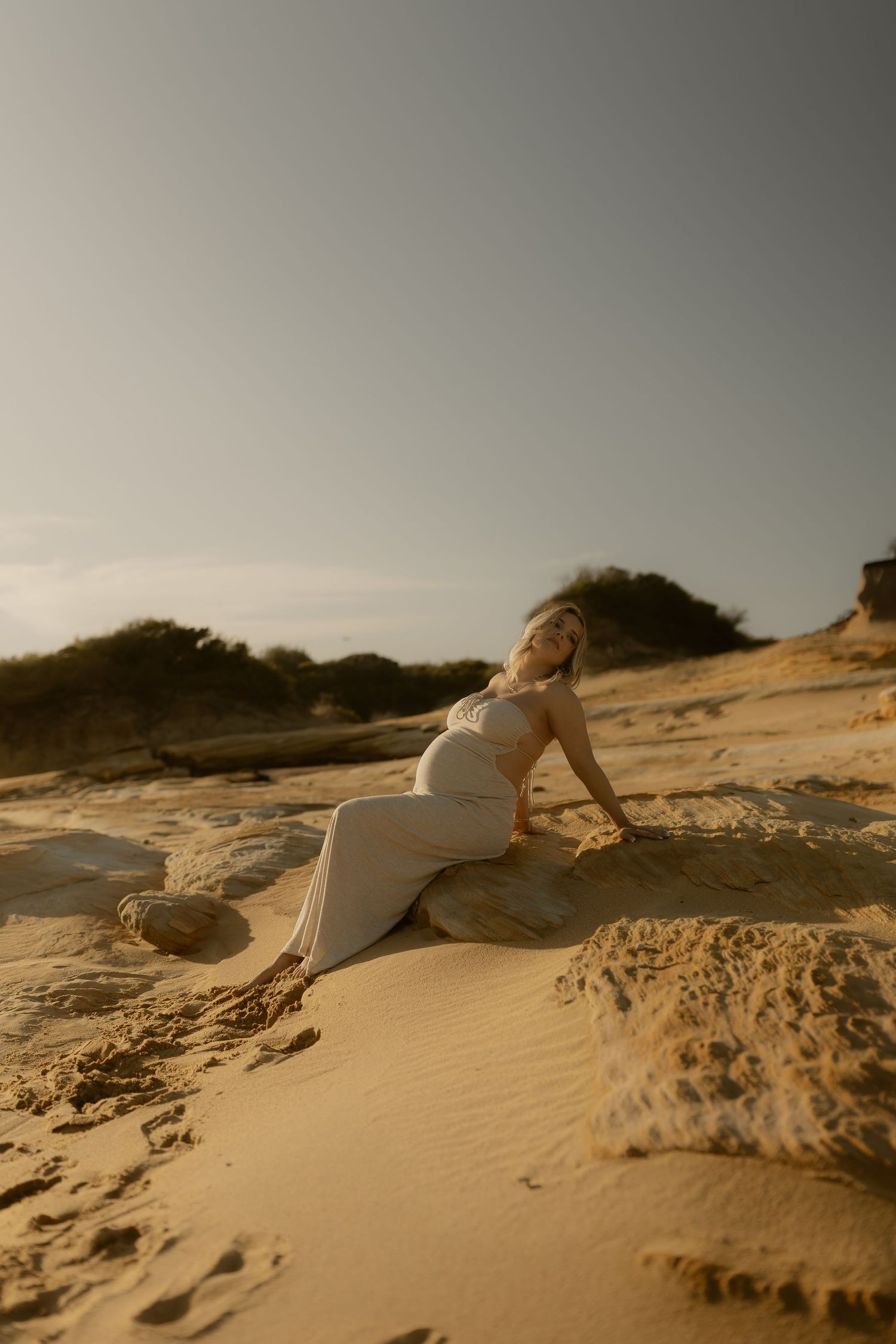 Figure in white dress lying gracefully on sandy dunes at sunset creating artistic silhouette against golden sky.