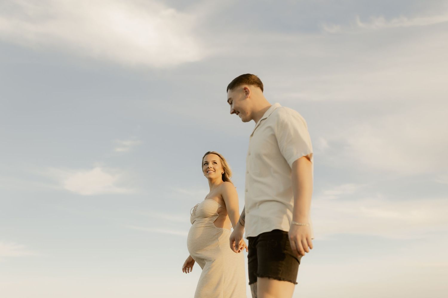 A couple stands together against a cloudy sky at sunset, with warm lighting creating a romantic silhouette.