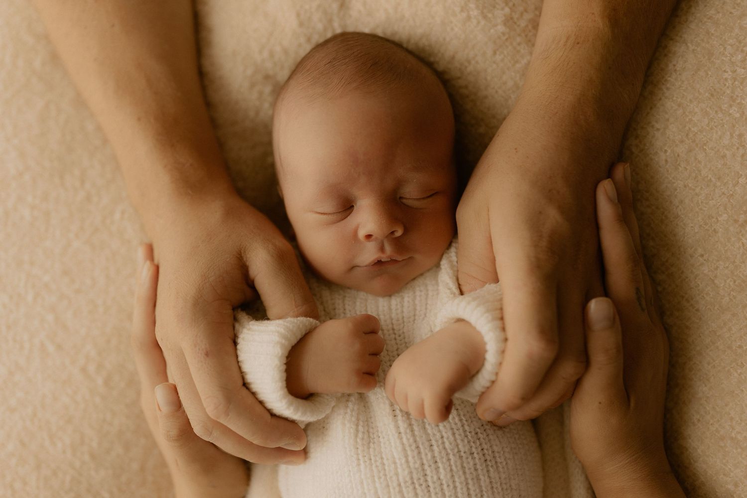 Gentle hands cradle a sleeping newborn baby wrapped in soft white fabric against a beige background.