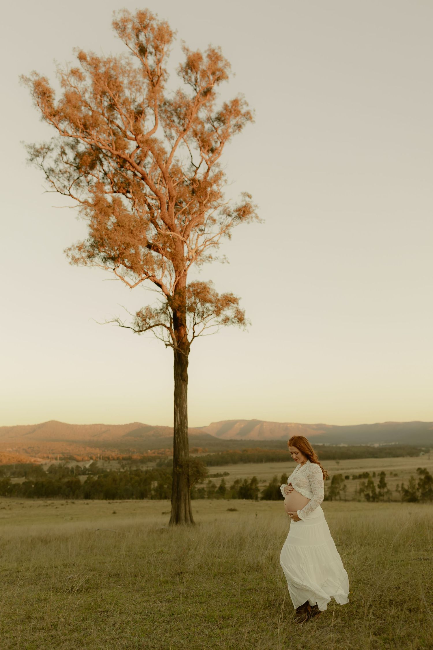 Pregnant woman in white dress stands beside tall tree at golden hour with mountain landscape backdrop.